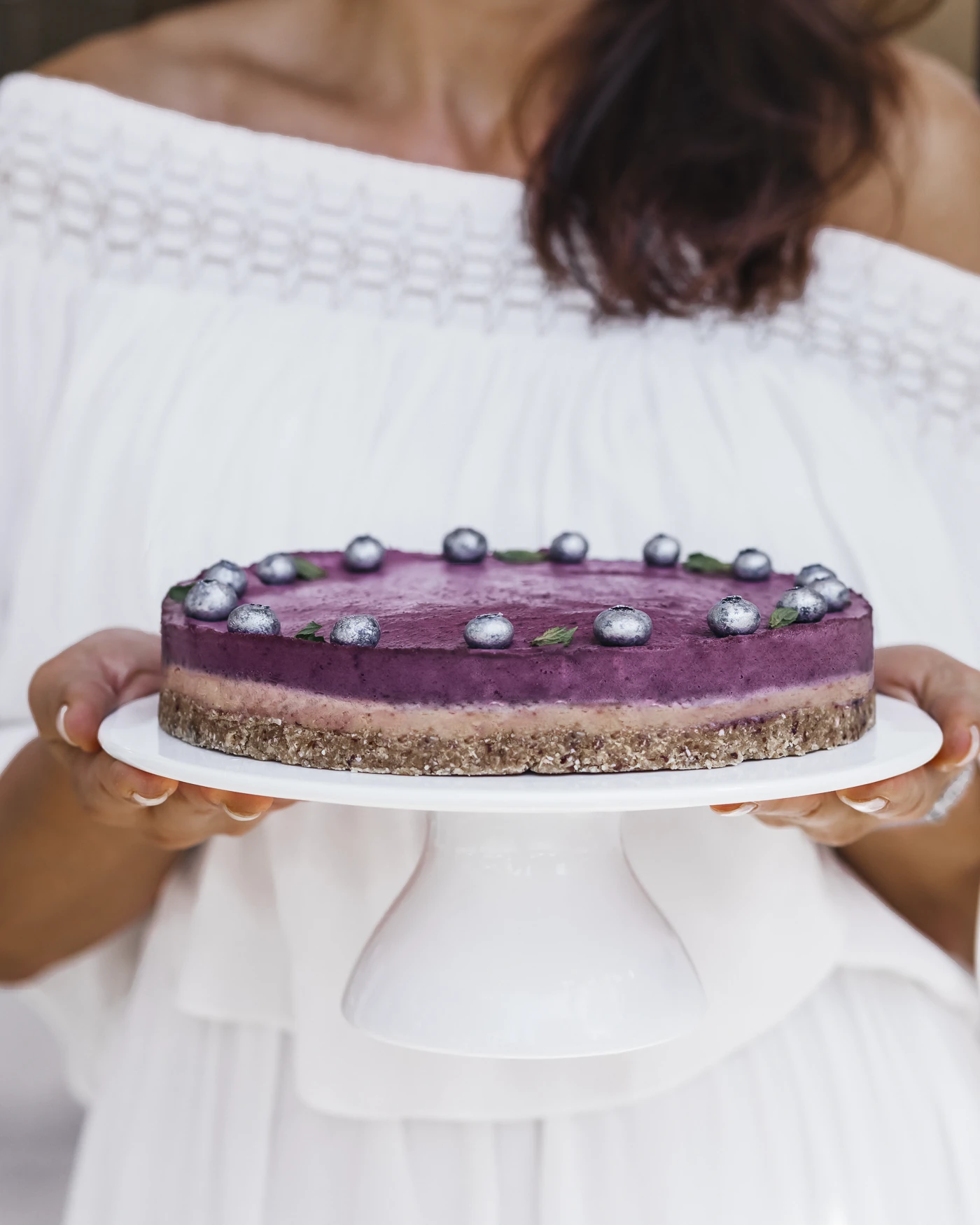 Lifestyle - woman presenting blueberry cake on pedestal, warm outdoor setting