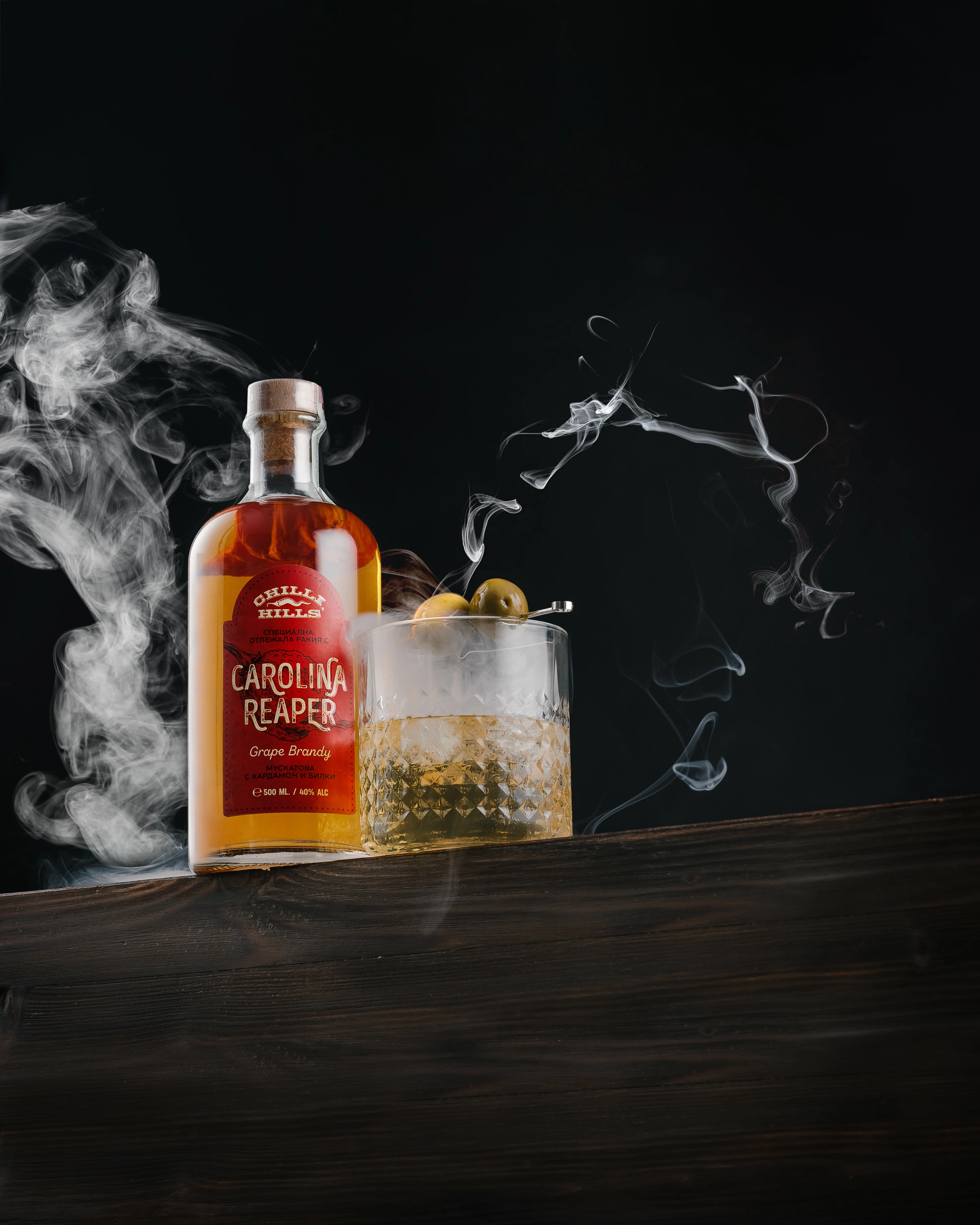 Chilli Hills Rakia Carolina Reaper Grape Brandy bottle with dramatic smoke and cocktail glass, dark background