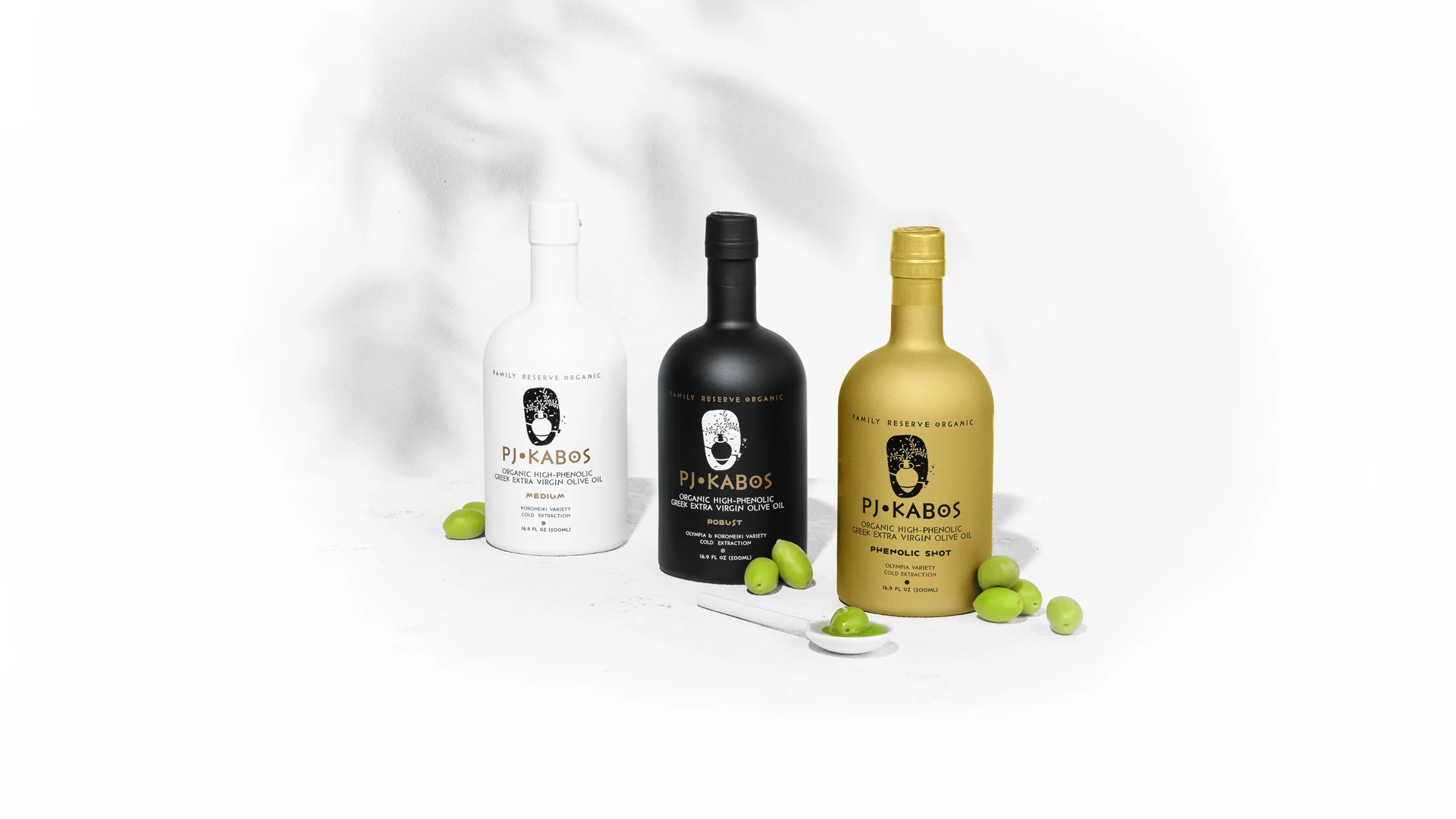 Премиум бутилки и тенекии PJ KABOS, подредени със средиземноморски елементи, зелени маслини, маслинови клонки и естествена светлина върху златистия зехтин