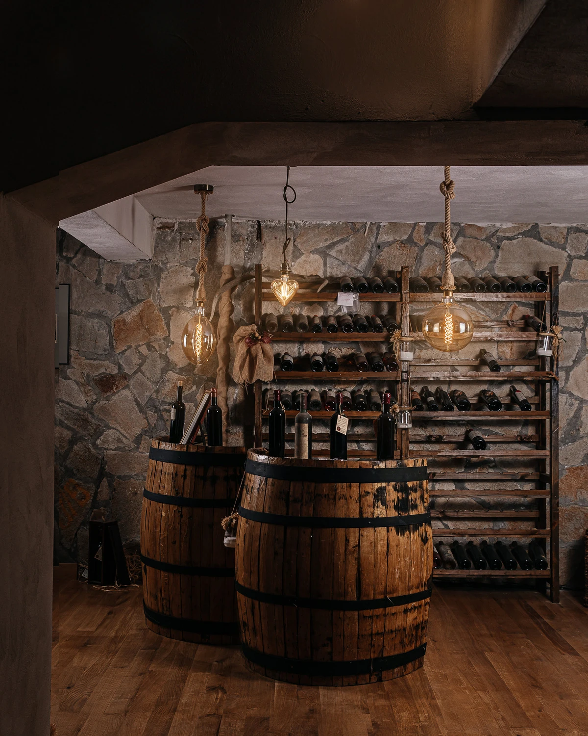 Винарна Вила Винифера: рустикална каменна изба с винени бъчви, стелажи за бутилки и топло винтидж осветление до Пловдив, България.