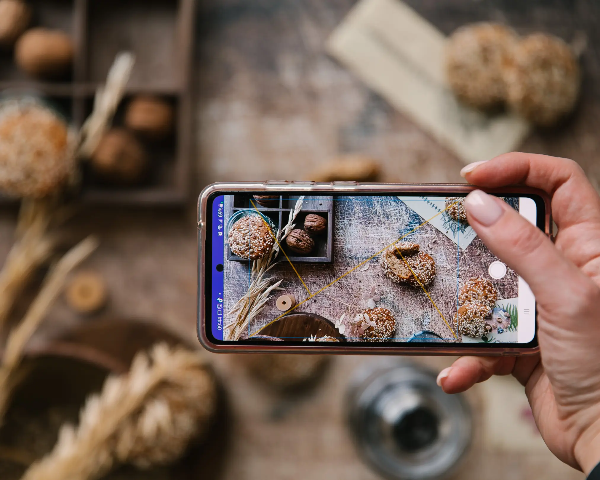 Ръка снима стилизирана храна: бисквити със семена и орехи, със смартфон с мрежа за триъгълна композиция.