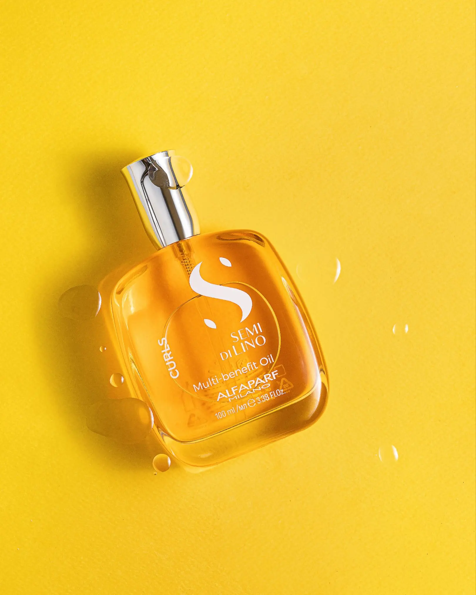 Semi di Lino Alfaparf Multi-benefit Curls Oil bottle with suspended droplets on a vibrant yellow background.