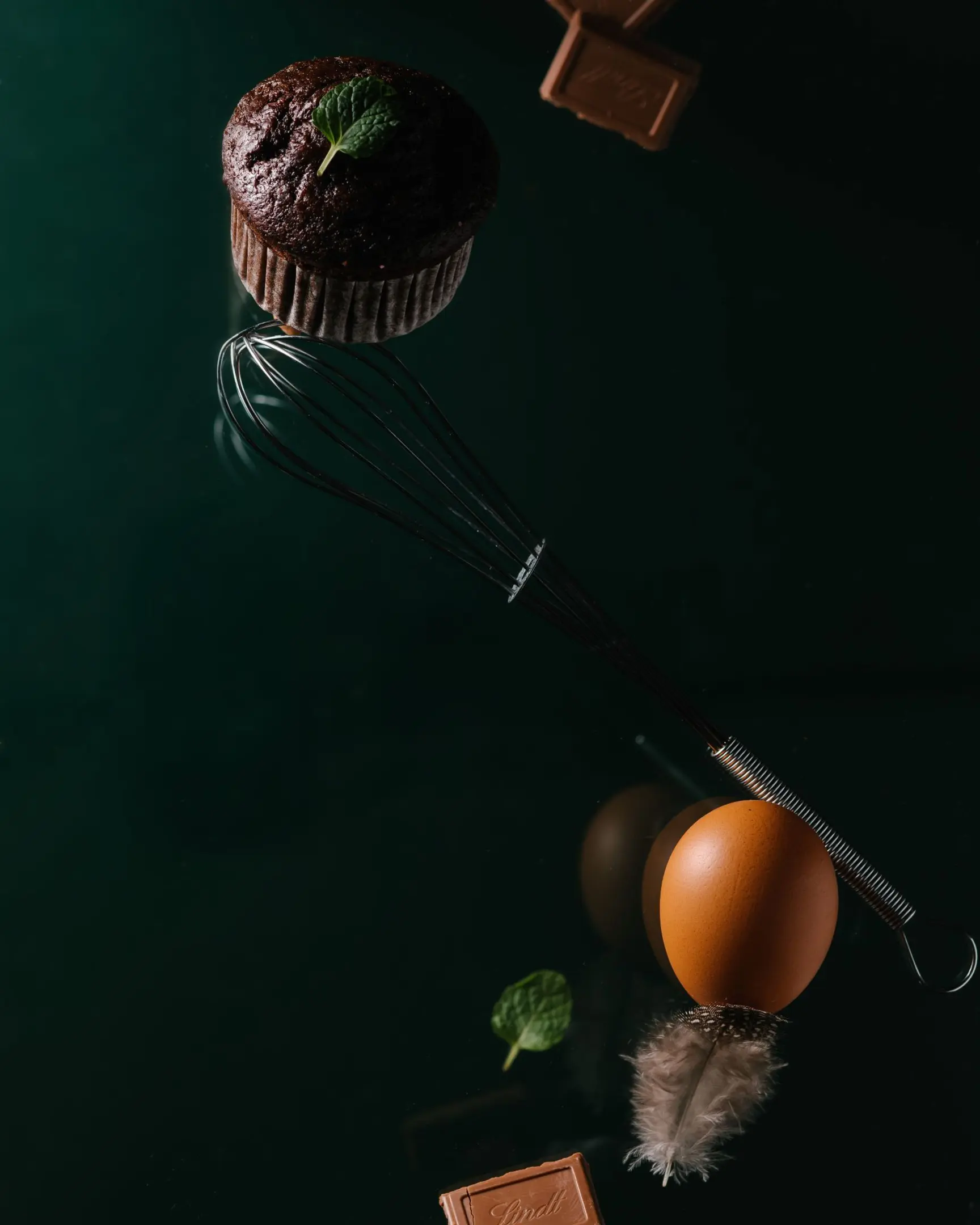 Dark chocolate cupcake with mint, whisk, egg on feather, Lindt chocolate floating on dark glass for product photography.