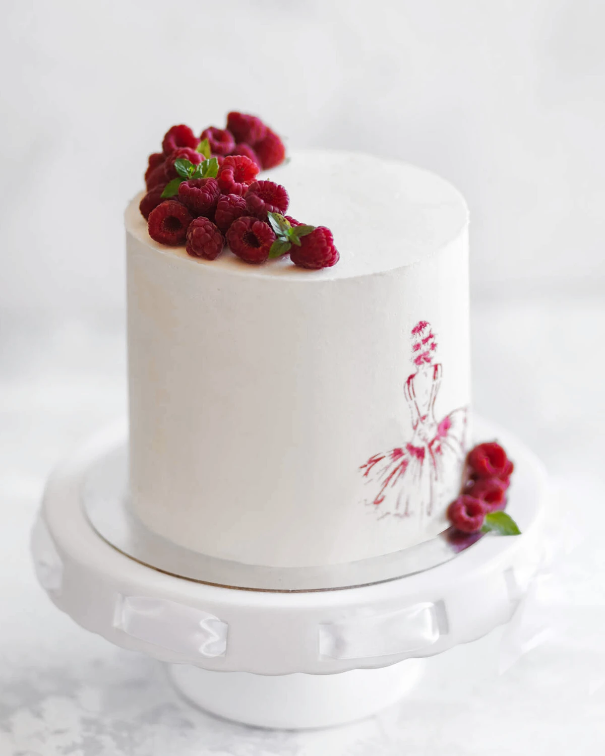 White sea buckthorn cake decorated with fresh raspberries and a red ballerina design, presented on a white cake stand.