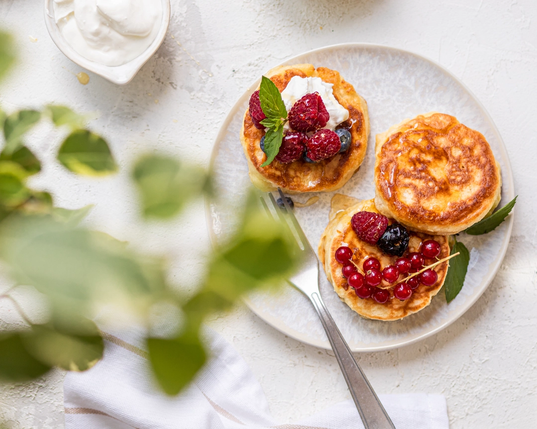 Golden fluffy kefir pancakes on a plate, topped with fresh raspberries, red currants, mint, and cream.