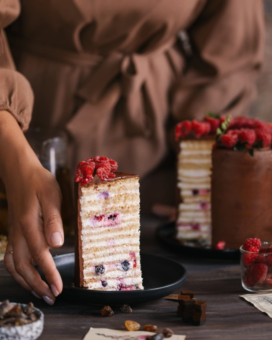 Hand holding a slice of Milky Girl milk cake with 14 condensed milk layers, berries, chocolate, and cream.