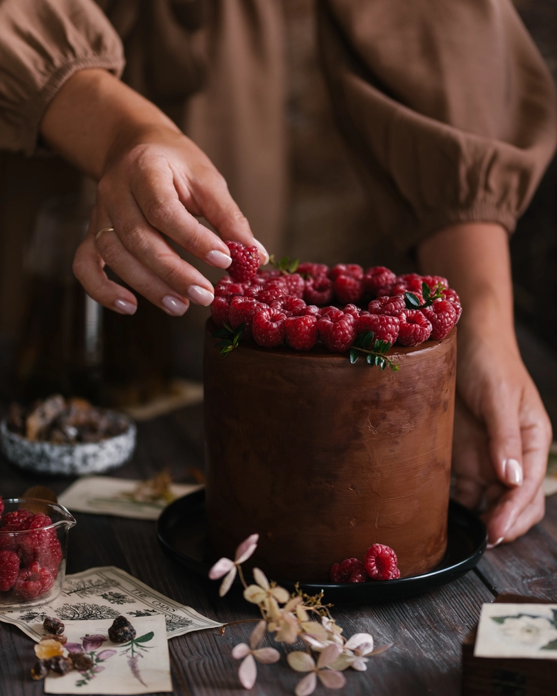 Hands decorating a tall Milky Girl chocolate cake with fresh raspberries on a dark wooden table.