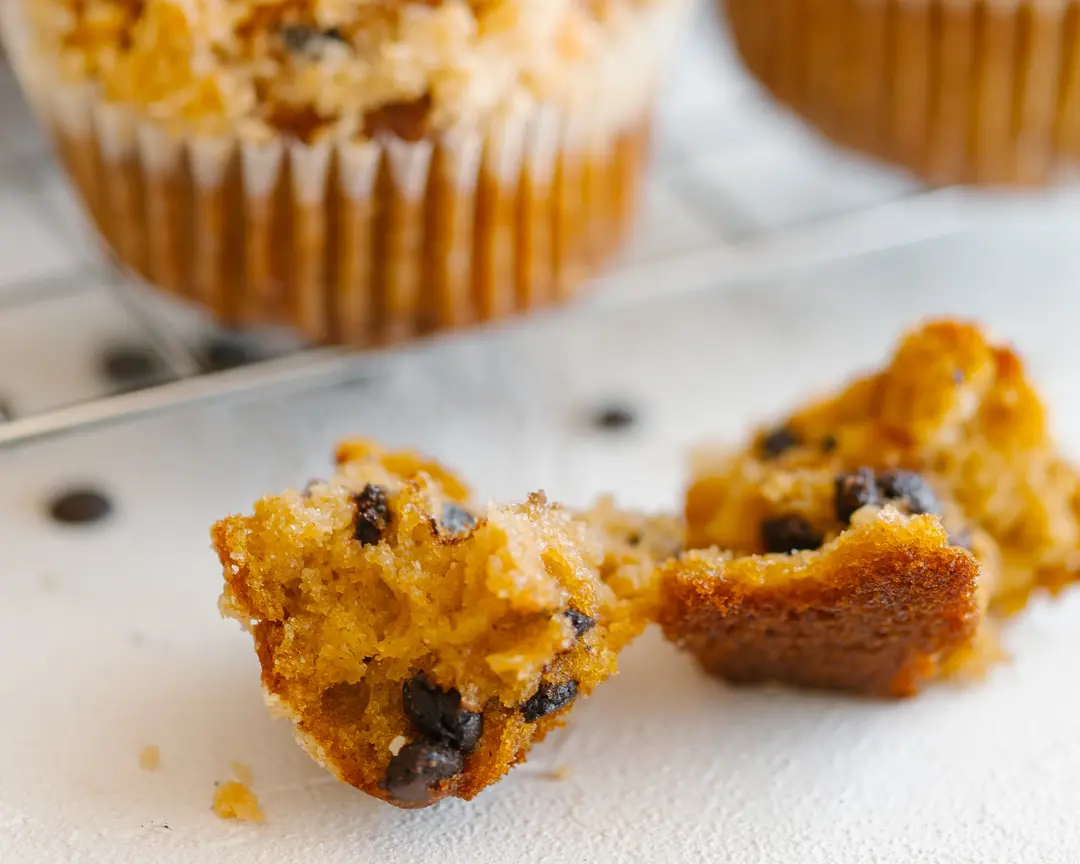 A freshly baked pumpkin muffin, broken open to show chocolate drops & crumble, with another muffin in the background.