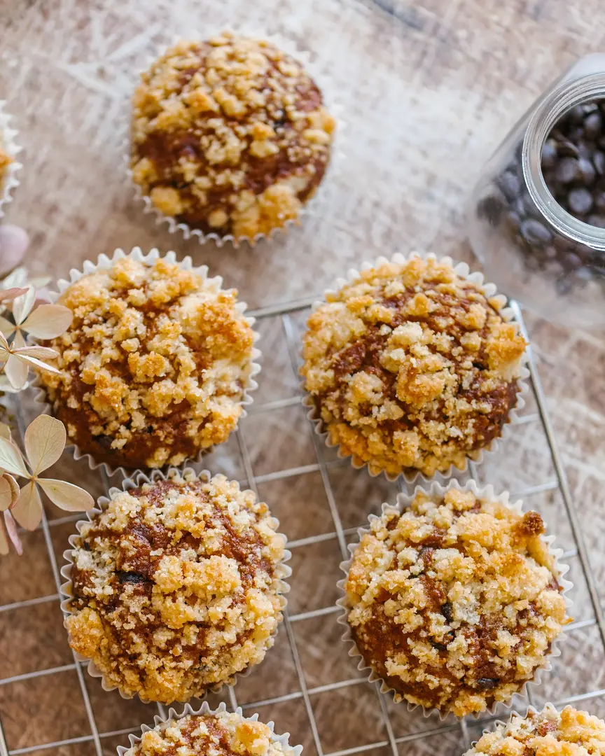 Freshly baked pumpkin muffins with crumble topping cooling on a rack, with a jar of chocolate drops nearby.