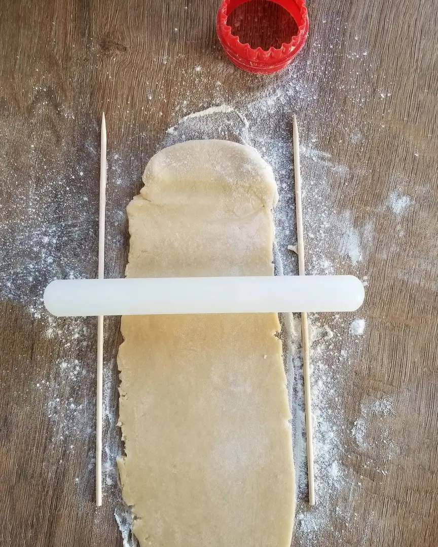 Разточване на тесто за бисквити равномерно с точилка, между дървени шишчета. Червена формичка за бисквити на набрашнена маса.