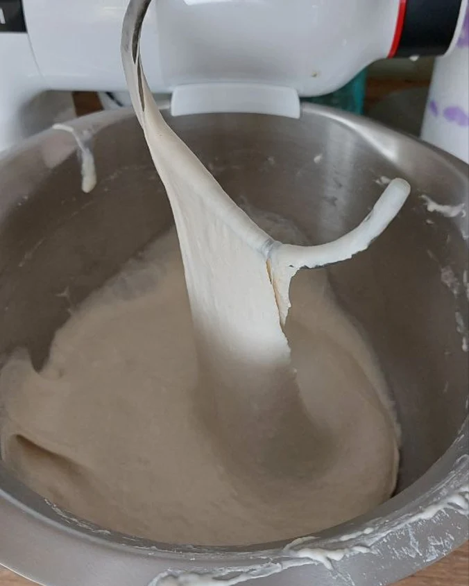 Кука за тесто на кухненски робот повдига гладко, лъскаво тесто за нощен хляб след последно омесване.