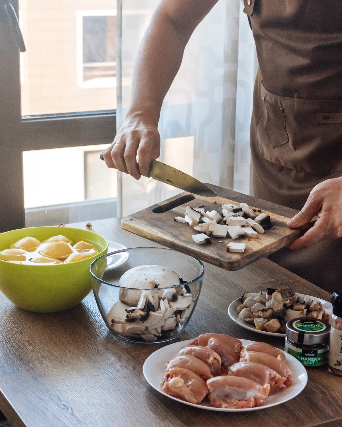 Person chops Portobello mushrooms for potato babka (kugel). Raw chicken, peeled potatoes, and Sharena Salt on kitchen counter.