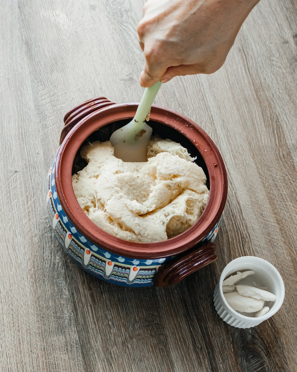 Hand preparing grated raw potatoes for traditional Belarusian potato babka (kugelis) in a decorative clay pot.