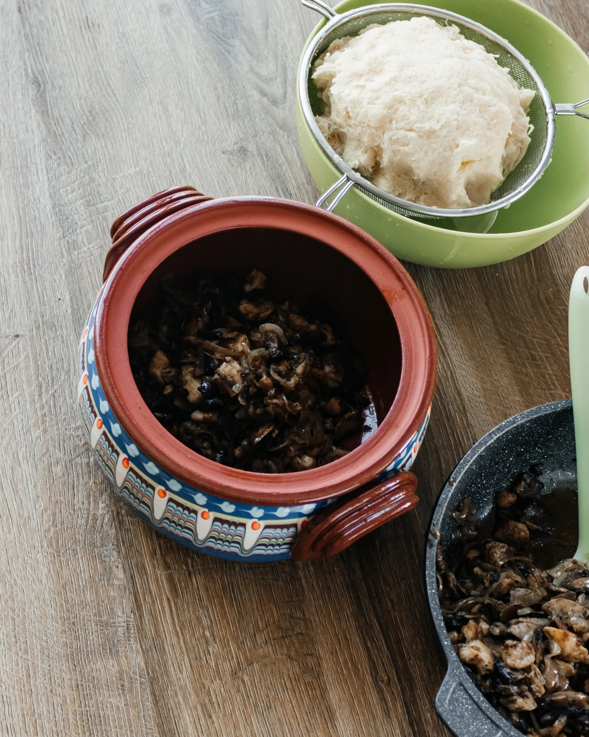 Grated raw potatoes in a strainer; fried mushrooms and onions in a clay pot and pan for Belarusian potato babka or kugel.