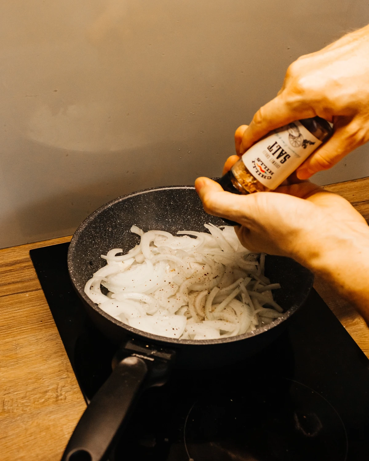 Hands add Chilly Hills Salt and pepper to sliced onions frying in a pan for Belarusian potato babka (kugelis).