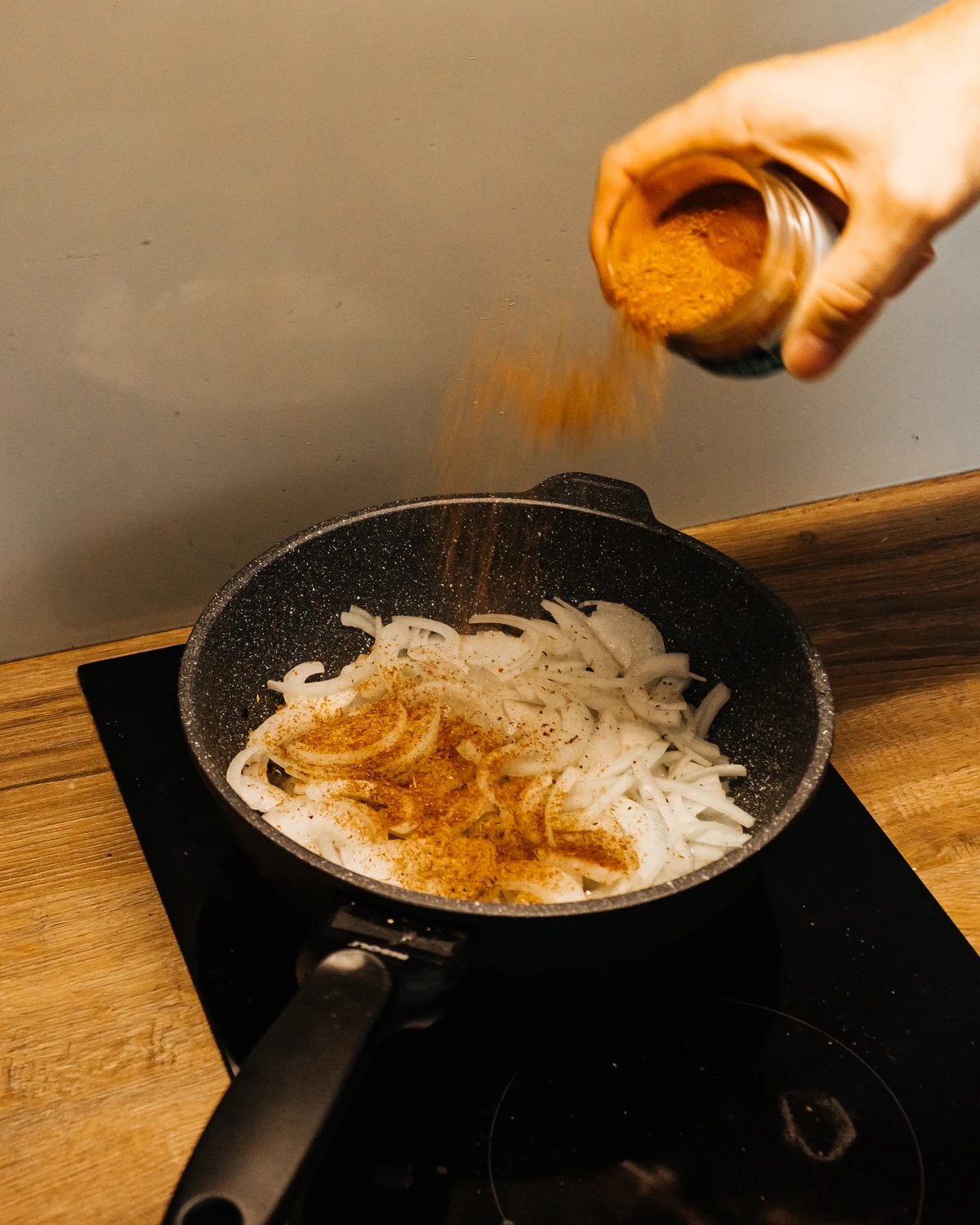 Person seasoning sliced onions in a frying pan, preparing ingredients for Belarusian Kugel or potato babka casserole.