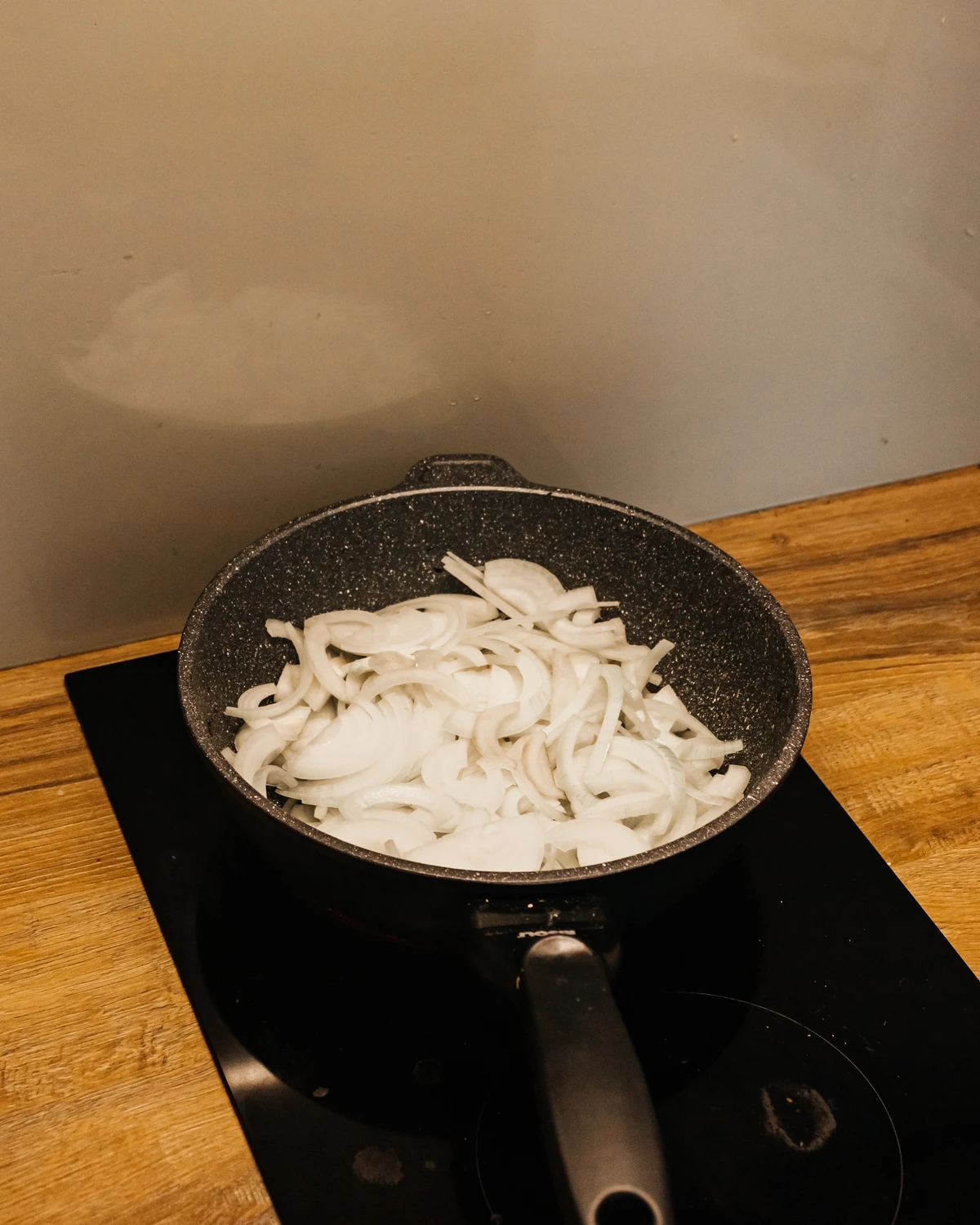 Sliced onions in a frying pan on an induction cooktop, a key ingredient for the potato babka or Jewish kugelis recipe.