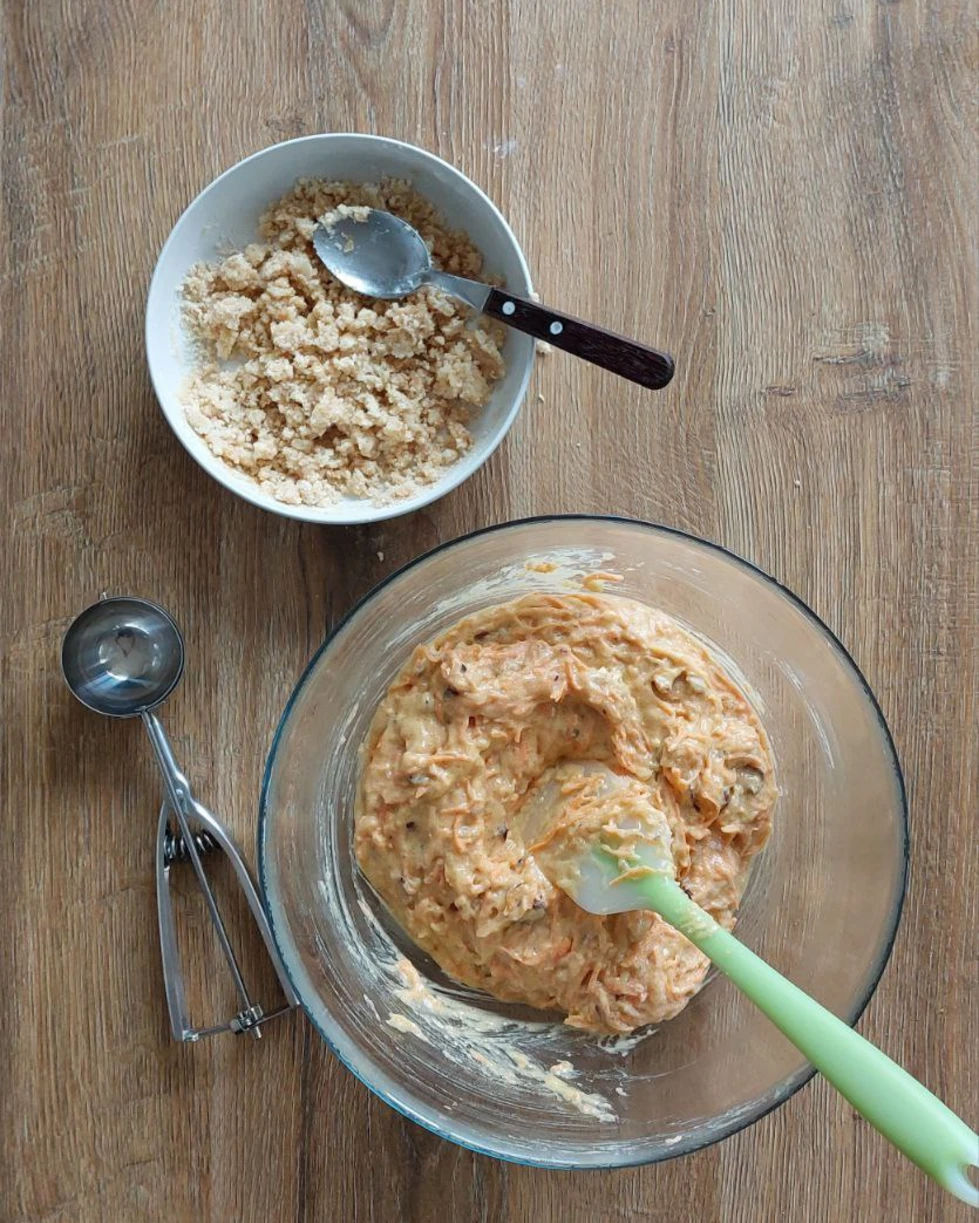Carrot muffin batter with a green spatula, a bowl of crumble topping, and an ice cream scoop for delicious homemade muffins.
