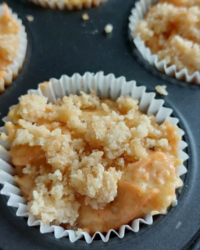 Unbaked carrot muffin batter topped with a generous, crunchy crumble in a muffin tin, ready to bake.