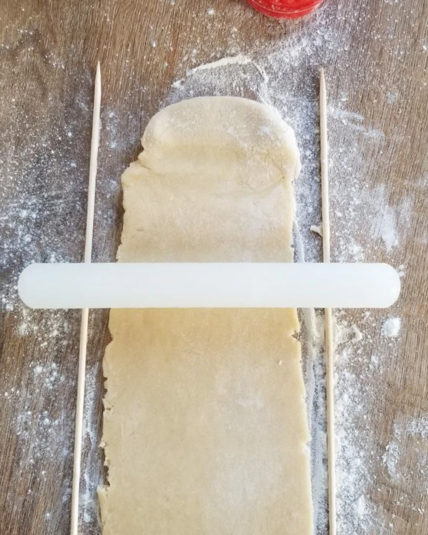 Rolling poppy seed shortbread cookie dough with a rolling pin between wooden skewers on a floured surface.