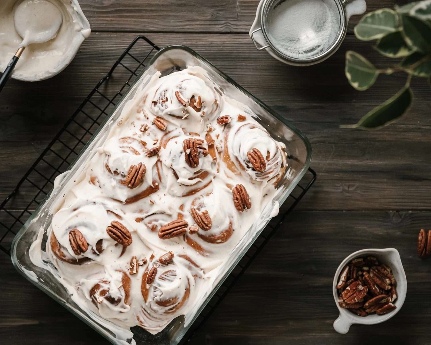Cinnamon rolls in a glass baking dish, generously topped with cream cheese glaze and pecans on a wood table.