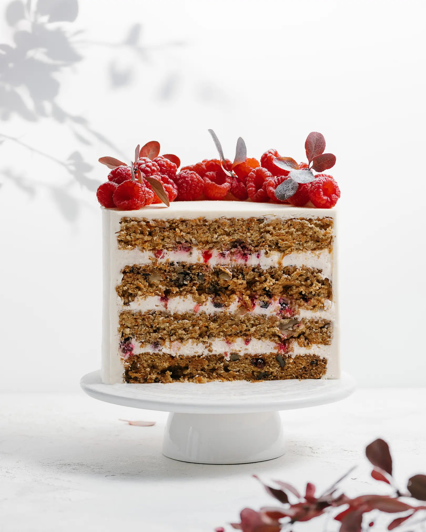 Multi-layered carrot cake on a stand, showcasing moist cake with walnuts, dried cranberries, mascarpone cream, & fresh raspberries.