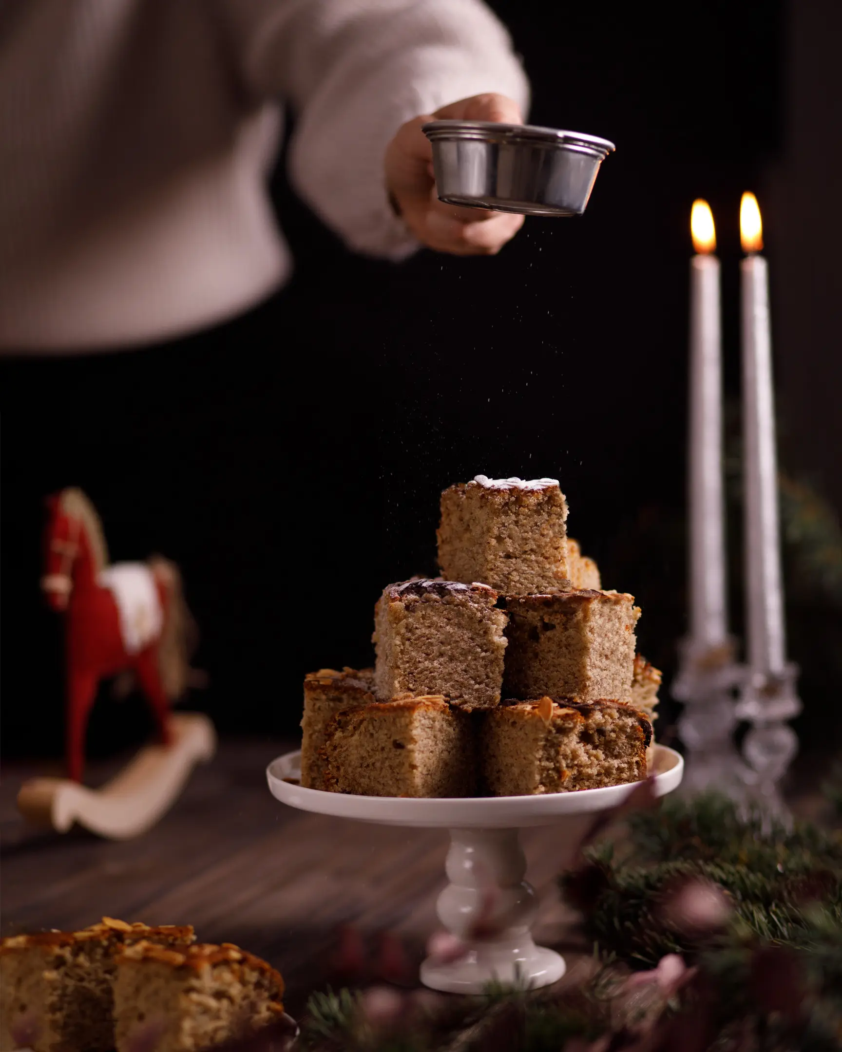 Ръка поръсва пудра захар над веган бананови квадратчета с бадемови люспи на празнична поставка за торта.