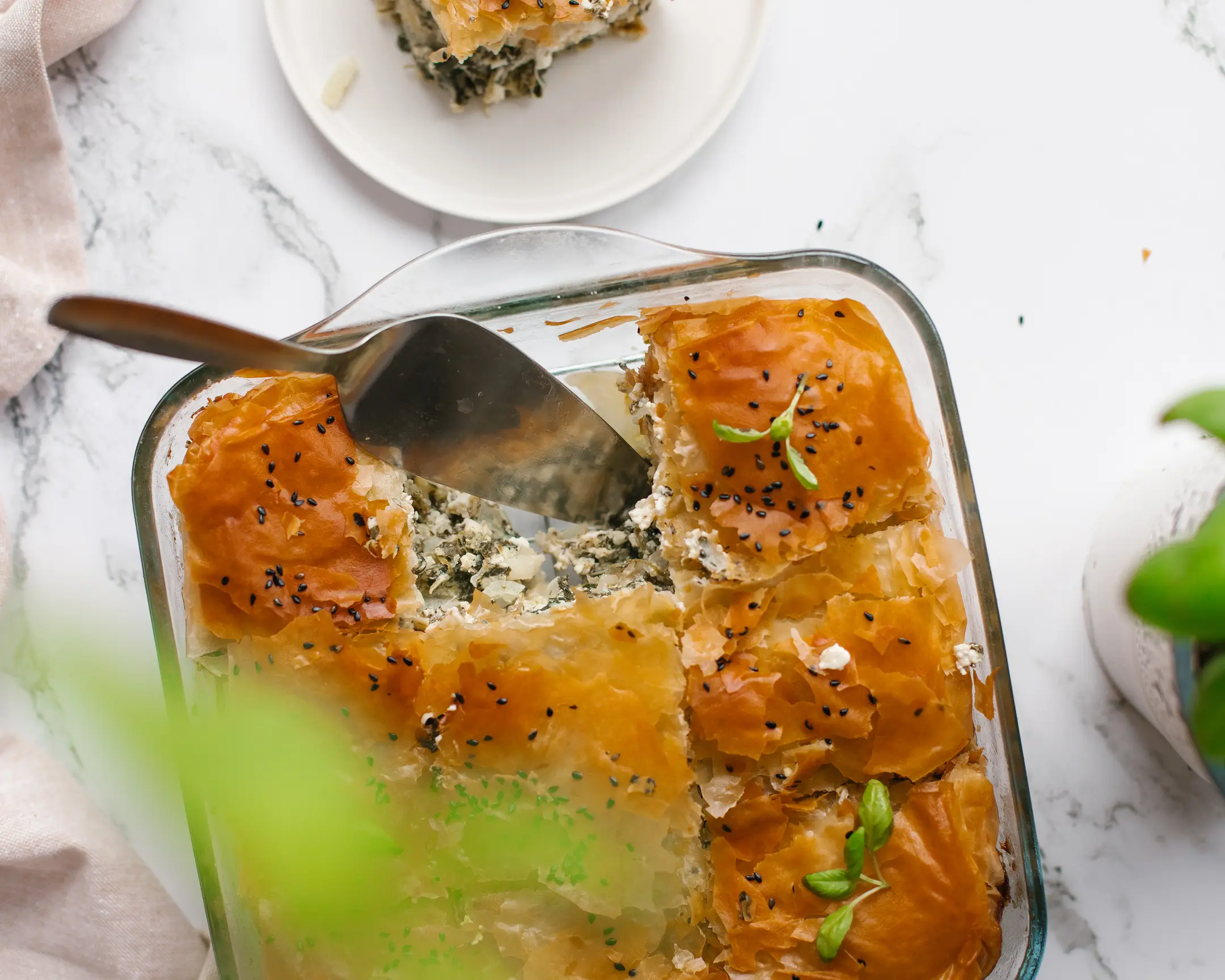 Top-down of baked spinach & feta spanakopita in a glass dish, a piece being served. Crispy filo pastry, Mediterranean recipe.