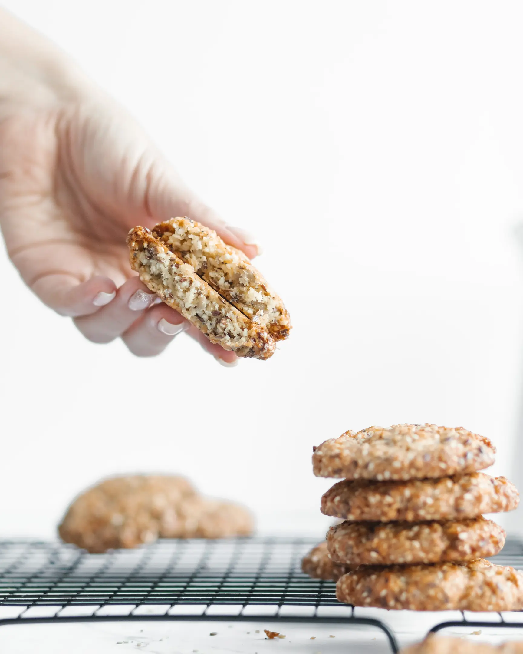 Woman's hand holds a split mixed seed cookie, revealing its texture. Freshly baked seed cookies stacked on a cooling rack.