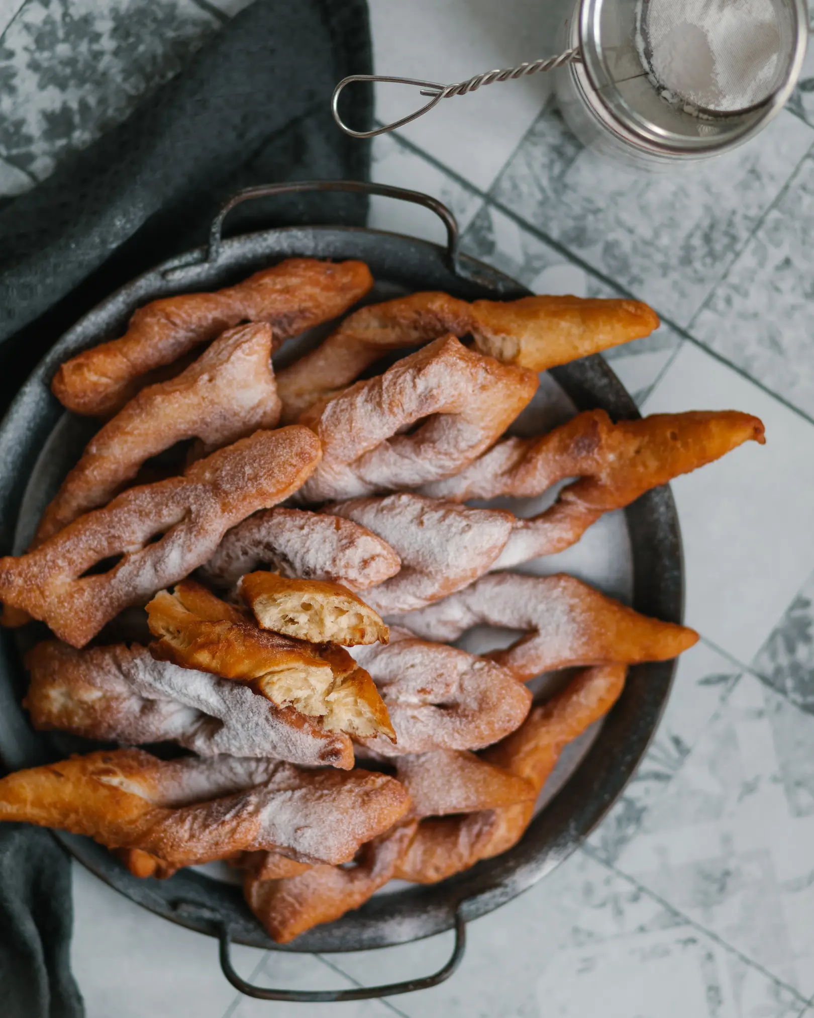 Crispy, golden Hvorost pastries on a tray, dusted with powdered sugar. One broken piece reveals light, fluffy texture.