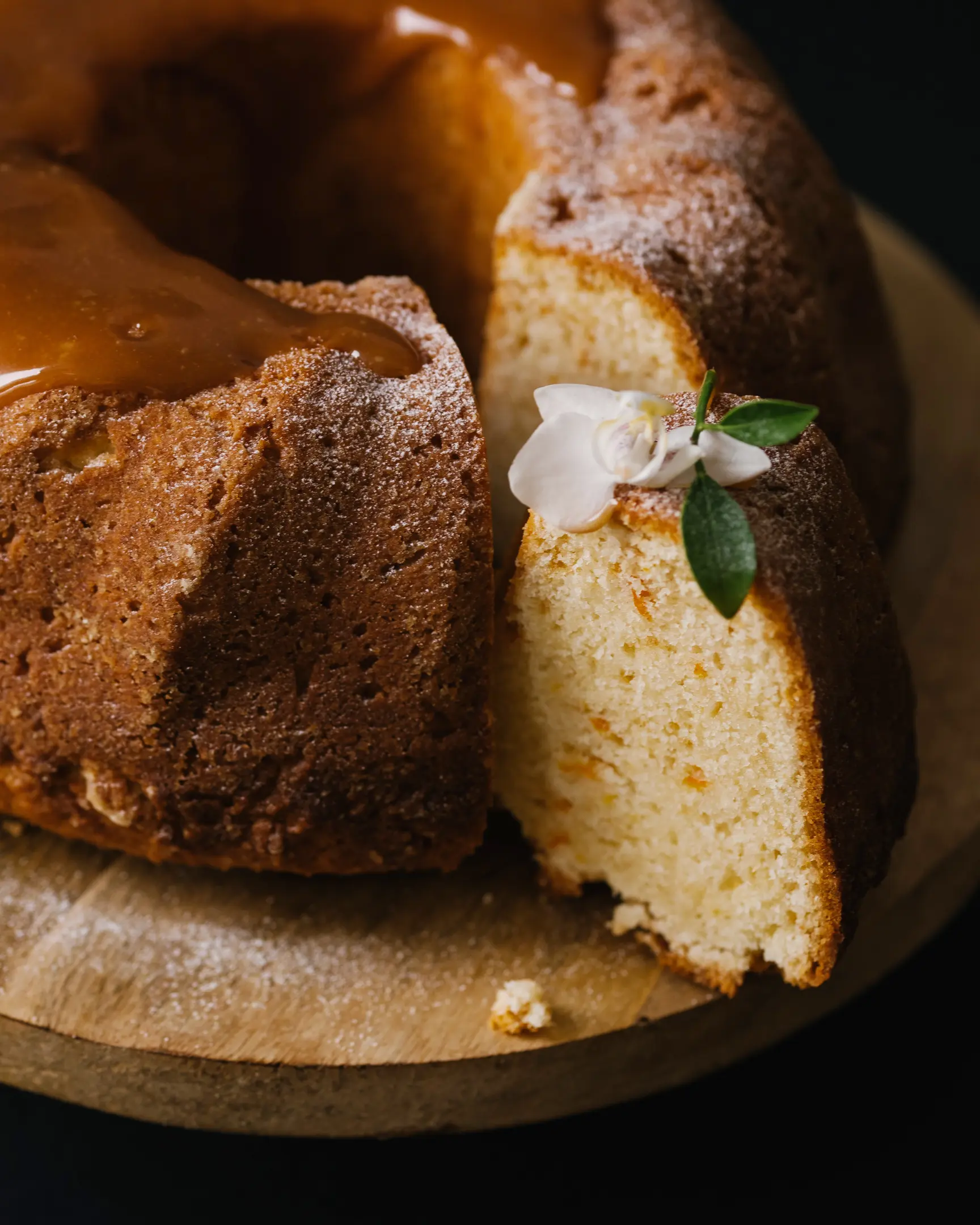 Beautiful bundt cake with caramel glaze, powdered sugar, and a slice showing orange zest, on a wooden cake stand.