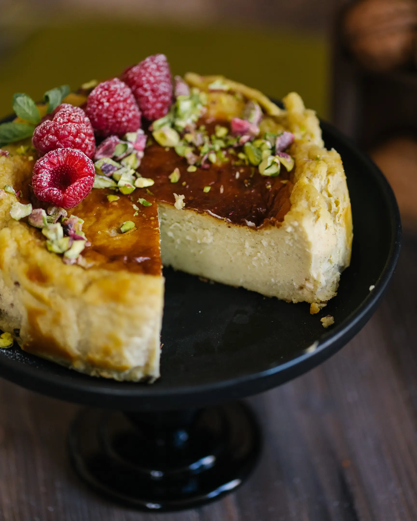 Pistachio Basque cheesecake with burnt top, creamy interior revealed by a slice, garnished with raspberries & pistachios.