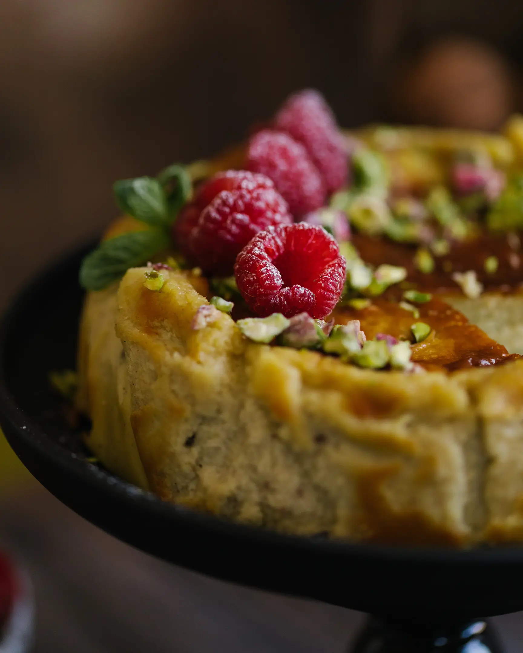 Whole pistachio Basque cheesecake, deep brown top, garnished with raspberries & pistachios, with a slice removed.