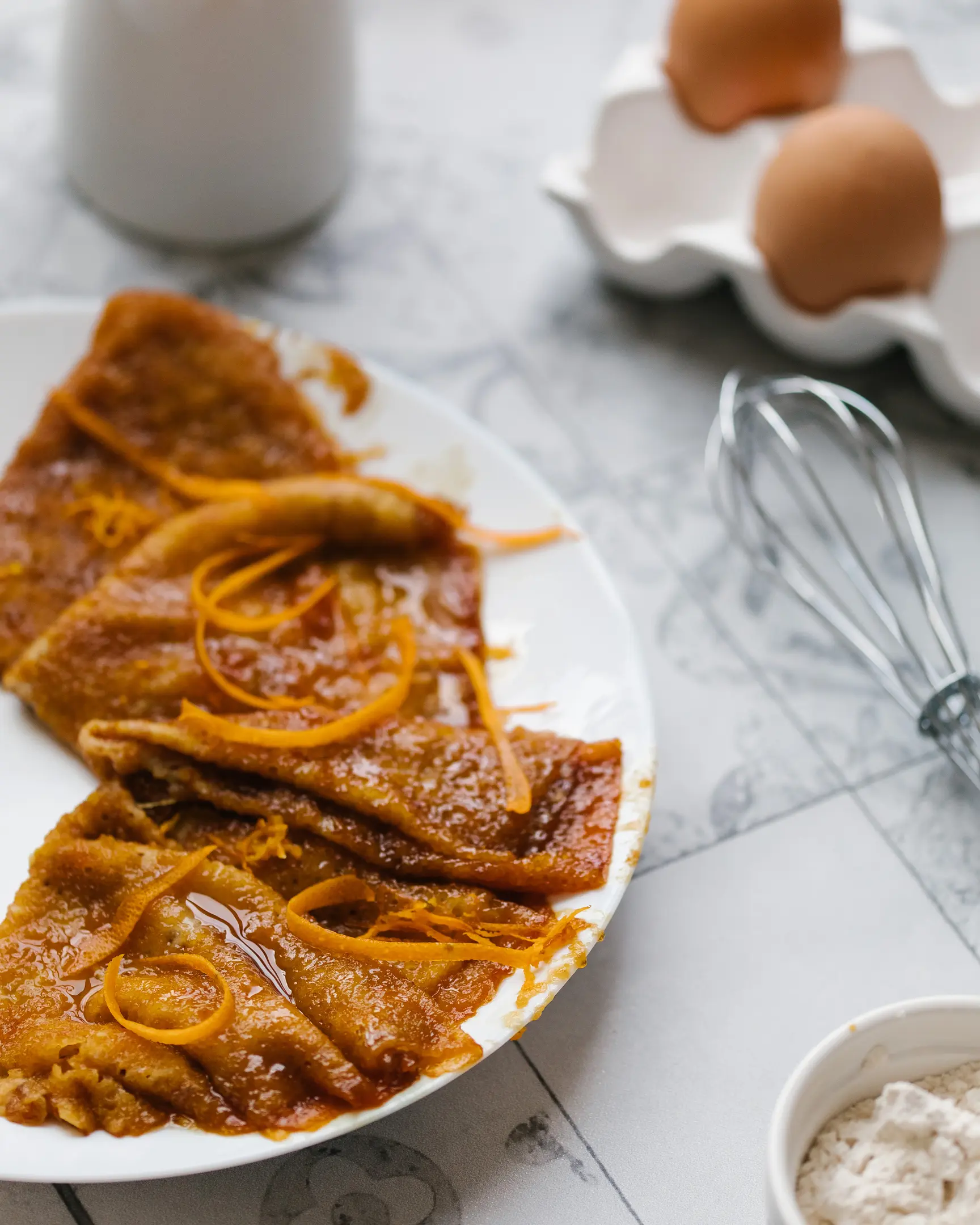 Златни палачинки с портокалов сос Коантро и корички. На заден план яйца, бъркалка, брашно за палачинки.