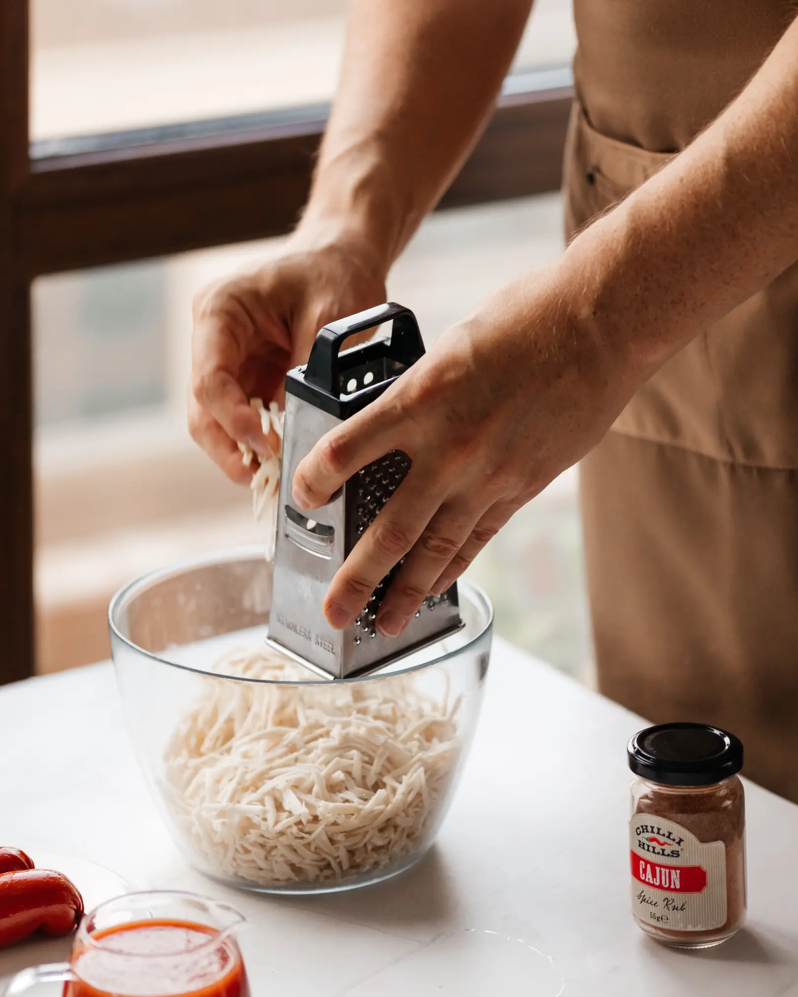 Съпруг настъргва сирене за домашна пица. CHILLI HILLS CAJUN подправка и доматен сос са готови.