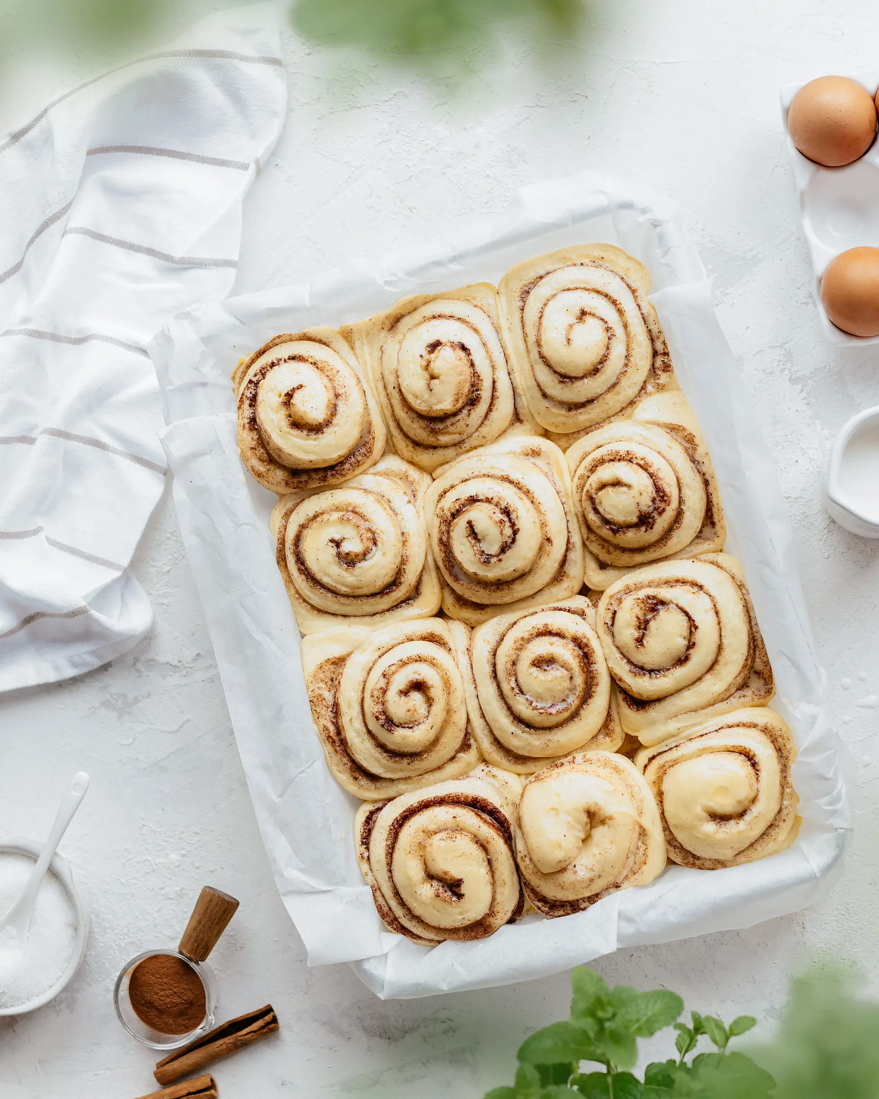 Неизпечени Cinnabon канелени рулца, втасващи в тава, заобиколени от яйца, мляко, захар и канела.