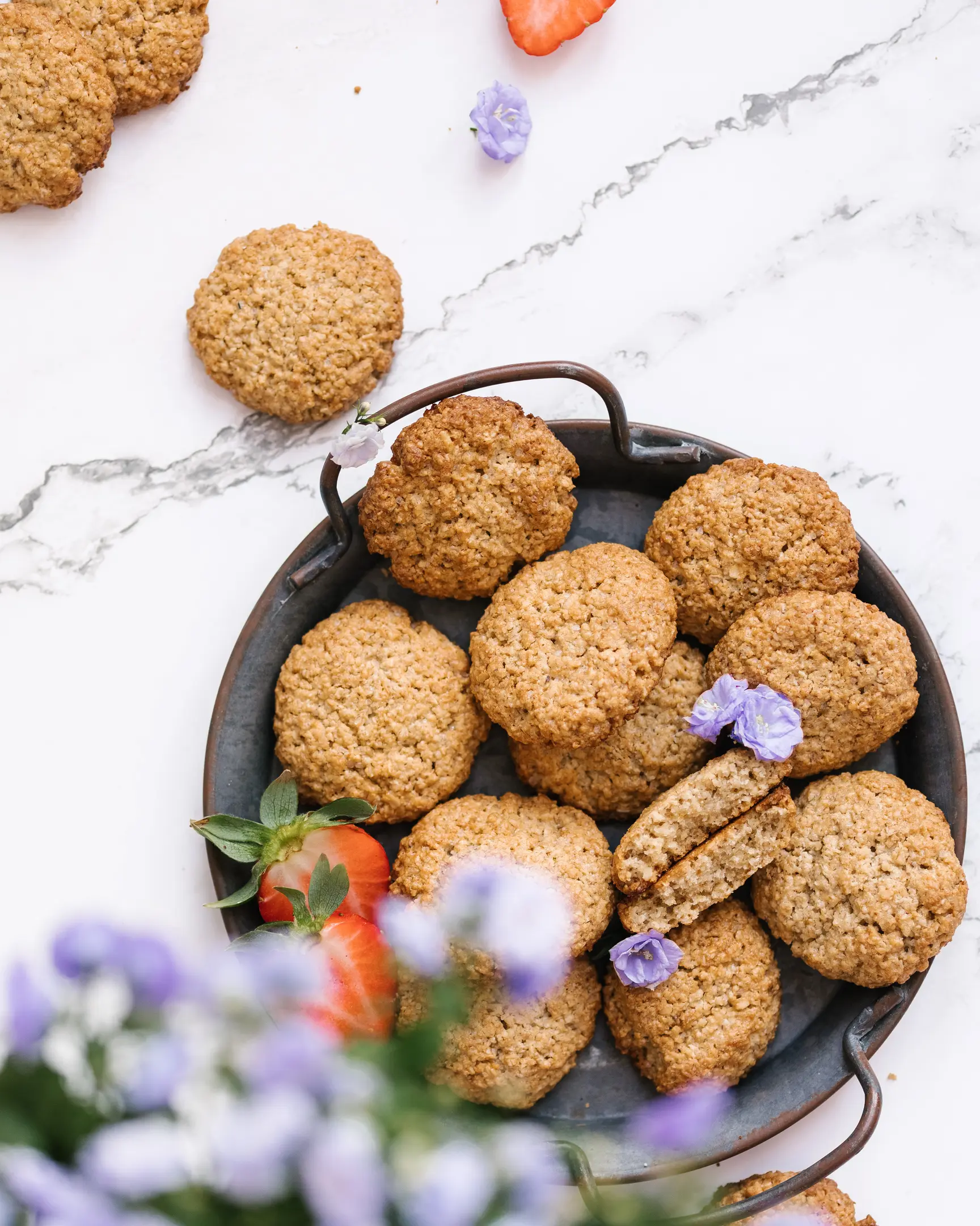 Oatmeal flourless cookies on a rustic tray with strawberries & purple flowers; one cookie broken to show texture.