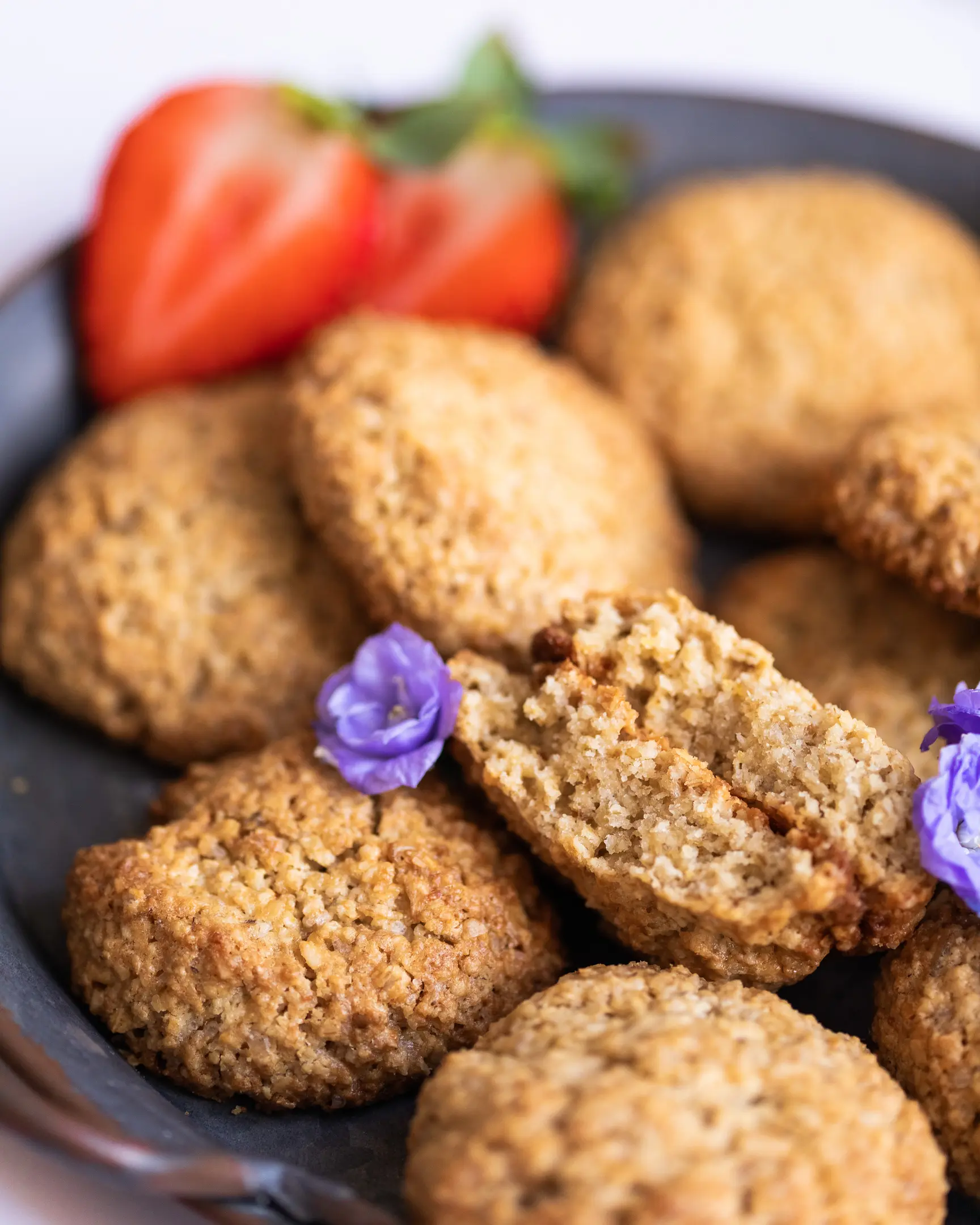 Freshly baked oatmeal flourless cookies, one halved to show texture, garnished with strawberries & purple flowers.