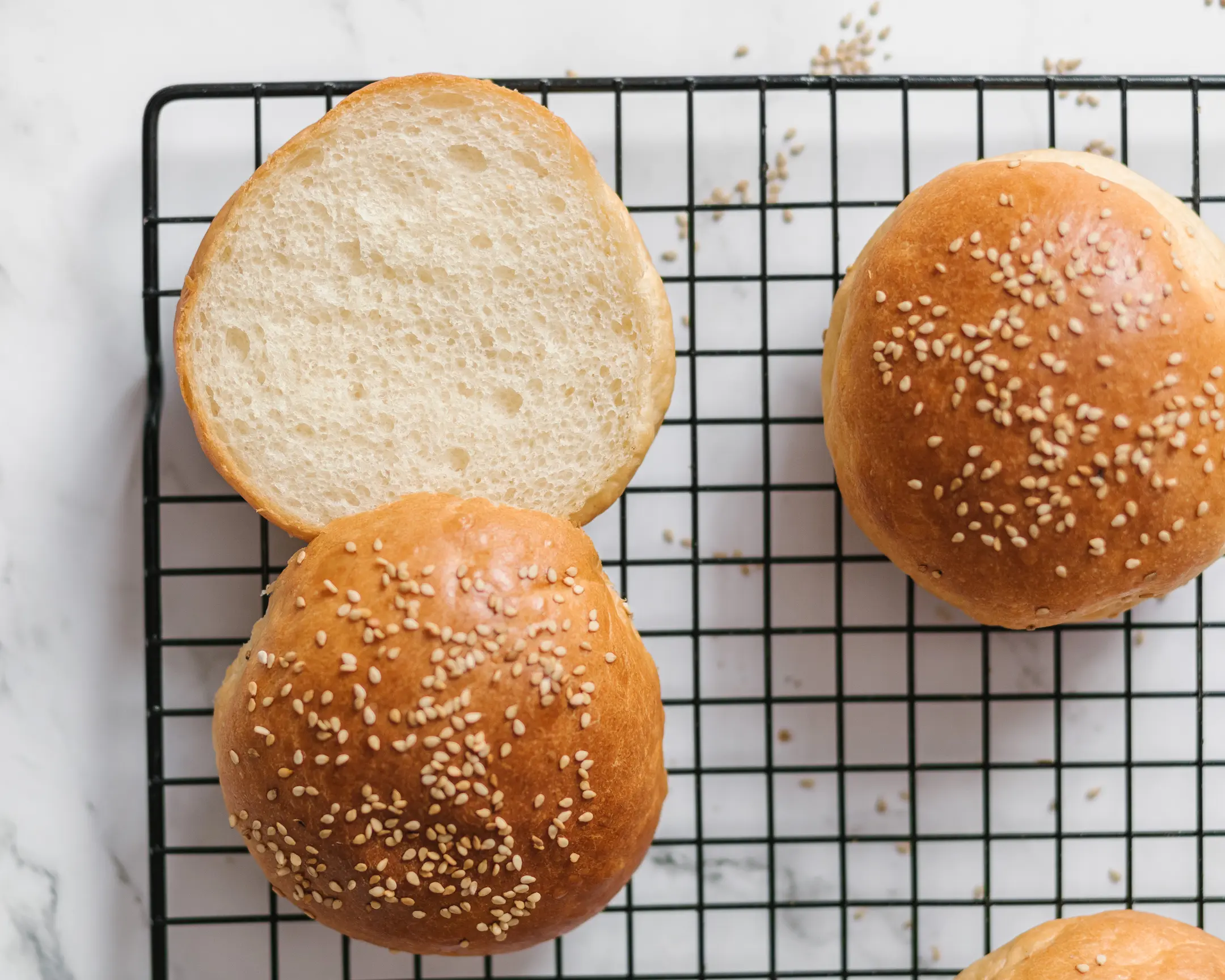 Soft, squishy potato burger buns for classic beef burgers, topped with sesame seeds, on a cooling rack; one bun is sliced.