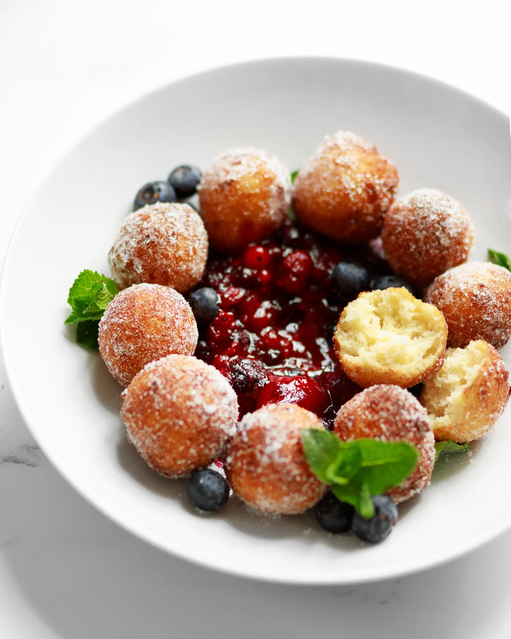 Golden bite-sized yeast-free cottage cheese donuts with powdered sugar, berry jam, blueberries, & fresh mint.