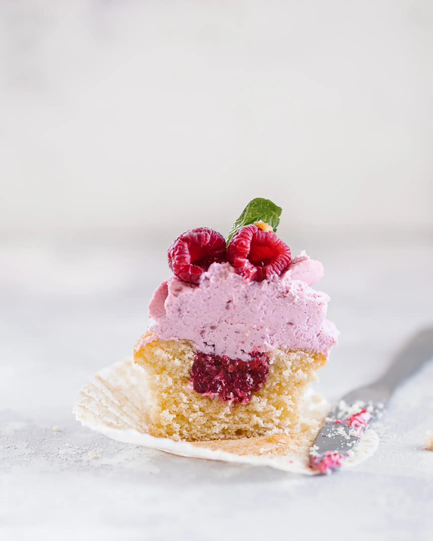 Cut vanilla cupcake revealing raspberry compote, topped with raspberry mascarpone cream, fresh raspberries & mint.
