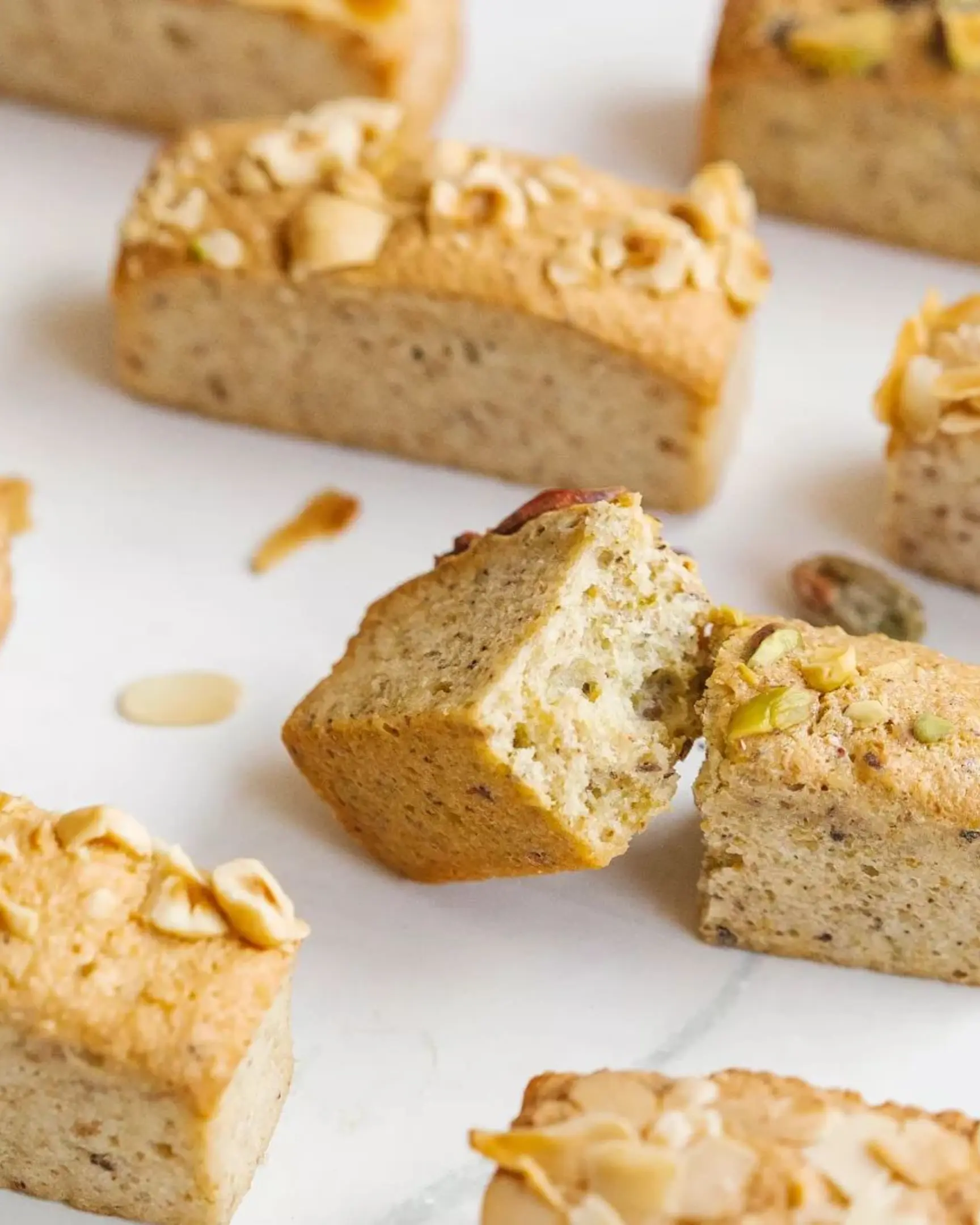 Freshly baked Financier pastries, gold bar shape, topped with nuts. One is cut open revealing its nutty interior.