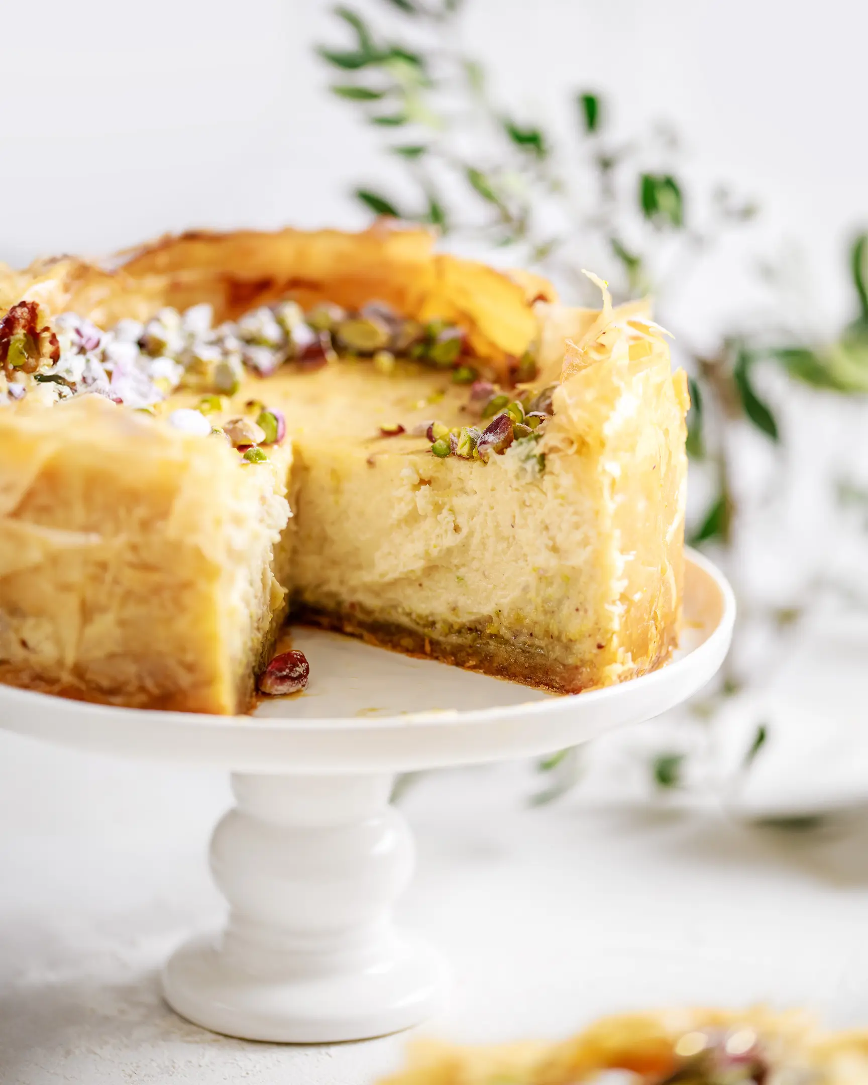 Ба клава чийзкейк на поставка, отрязано парче разкрива кремообразен пълнеж, слой шамфъстък и нагъната кора от фини кори.