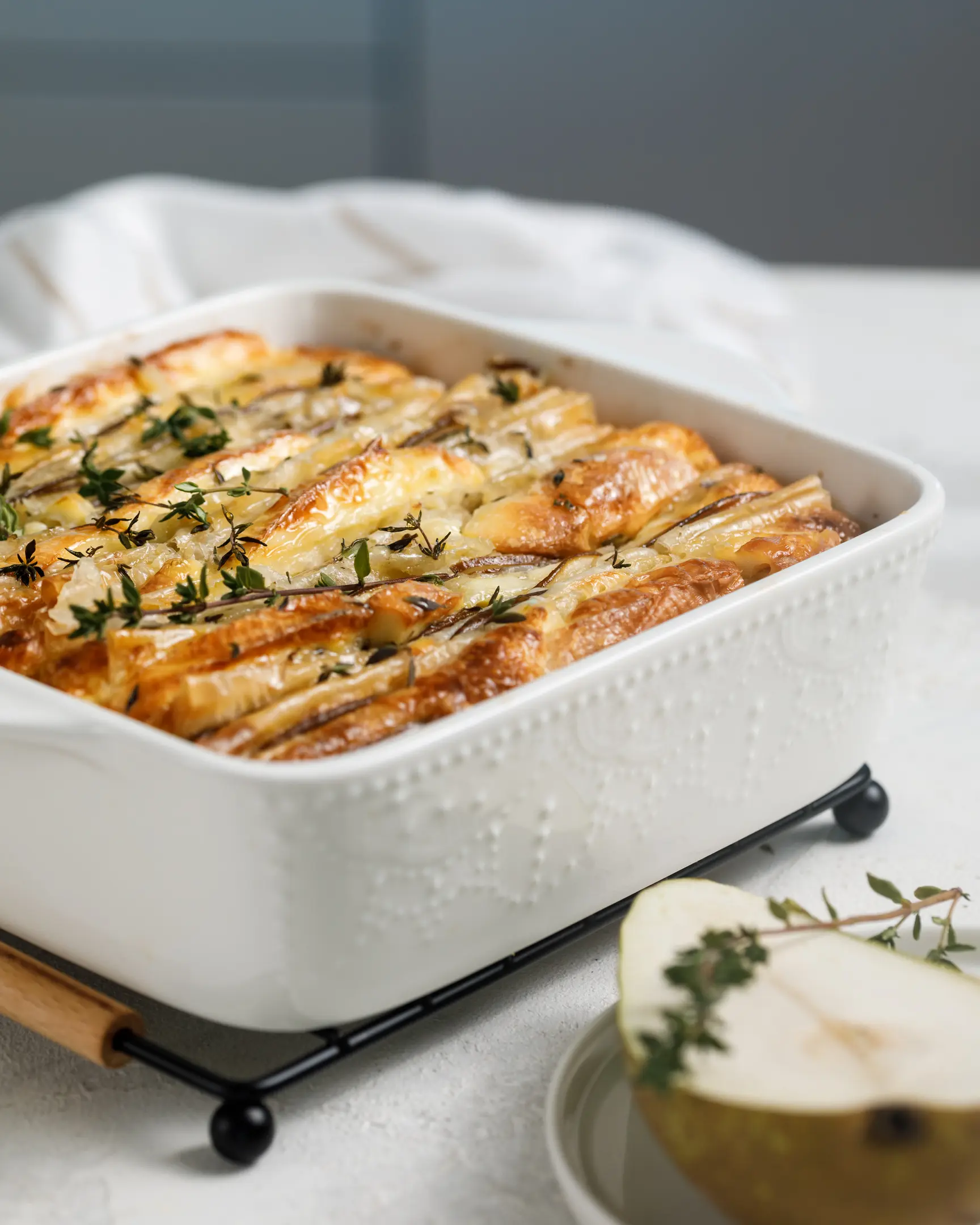Golden-baked Brie & pear filo tart topped with fresh thyme in a white dish, with a pear slice, perfect for dessert or brunch.