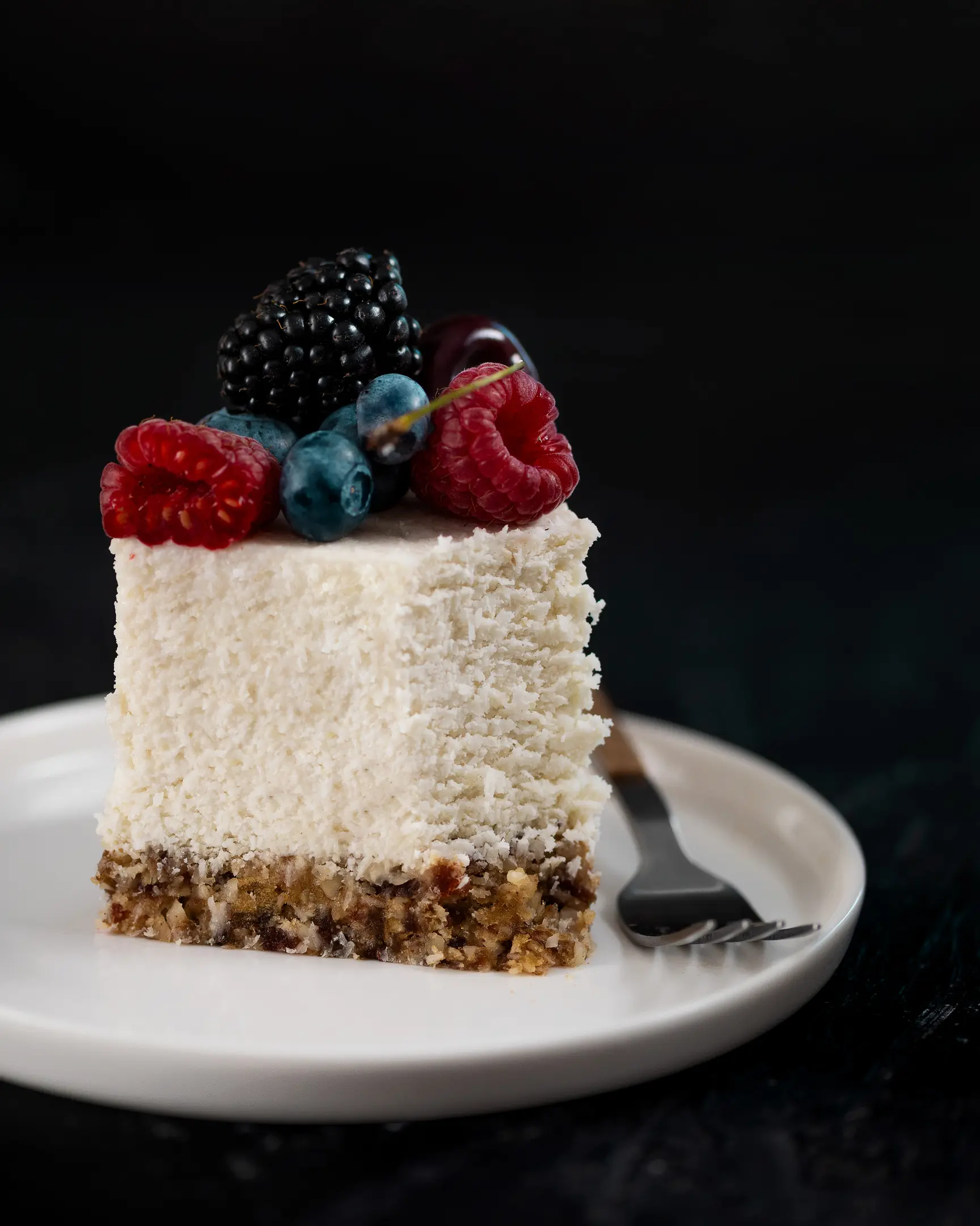 Creamy coconut raw cake slice, a tropical and guilt-free delight, garnished with fresh raspberries, blueberries, and blackberries.