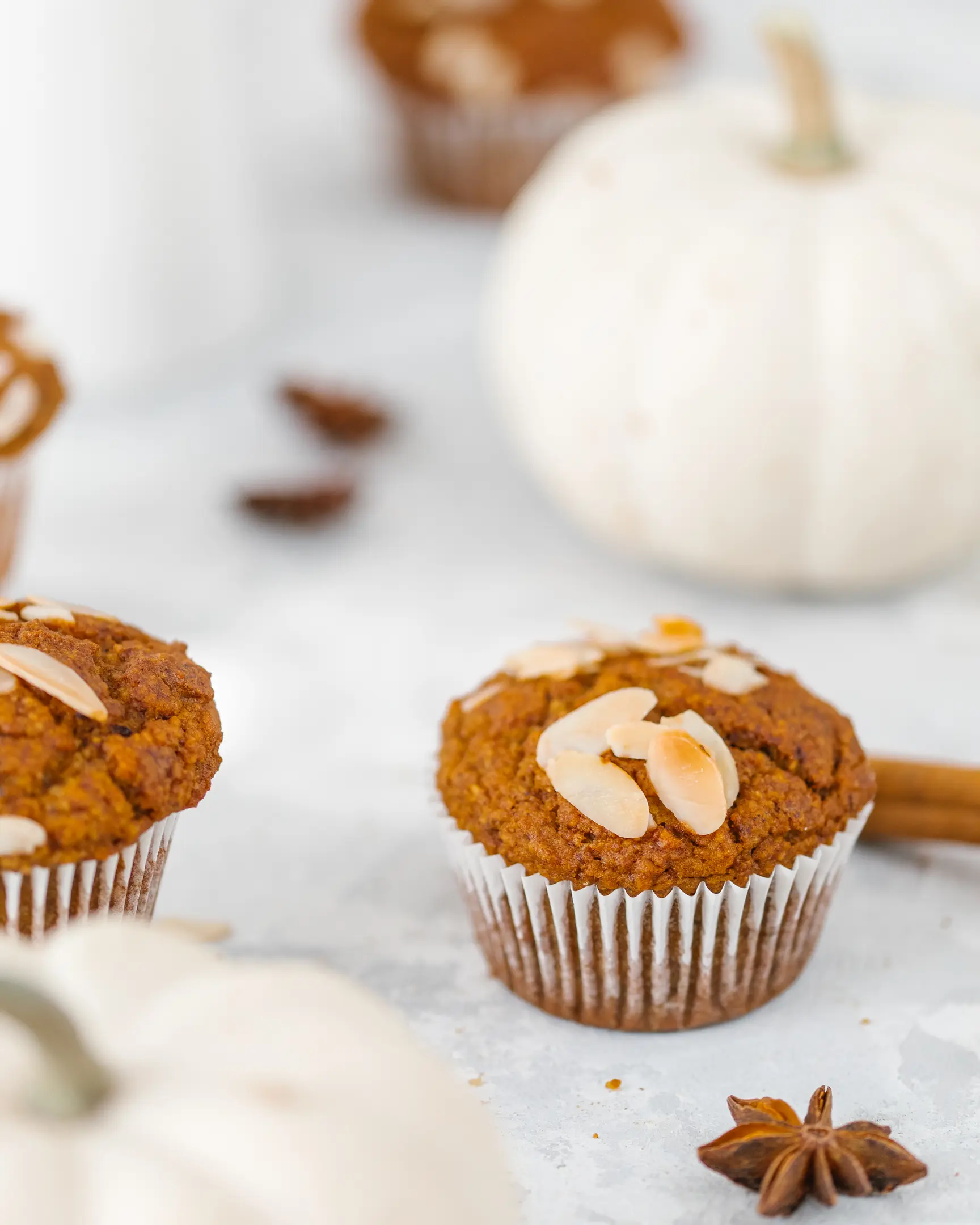 Moist pumpkin muffin topped with almond flakes, alongside cinnamon sticks and decorative white pumpkins.