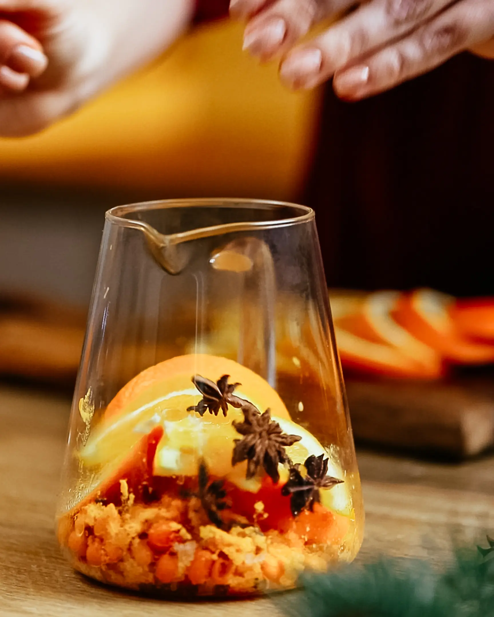 Adding orange slices, star anise, and mashed sea buckthorn berries to a glass pitcher for tea.