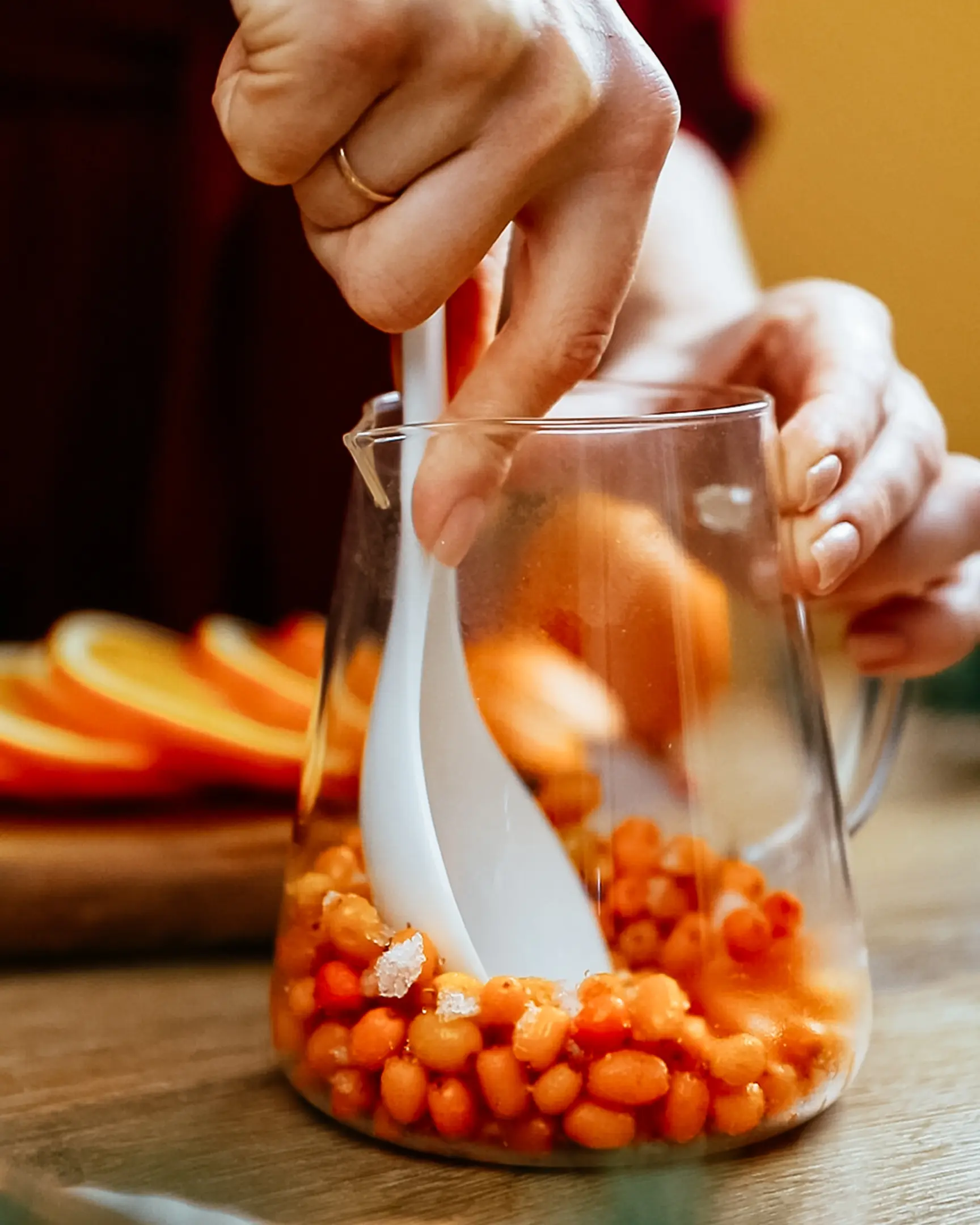 Hands mashing sea buckthorn berries in a pitcher with a spoon for tea; orange slices on wood board.
