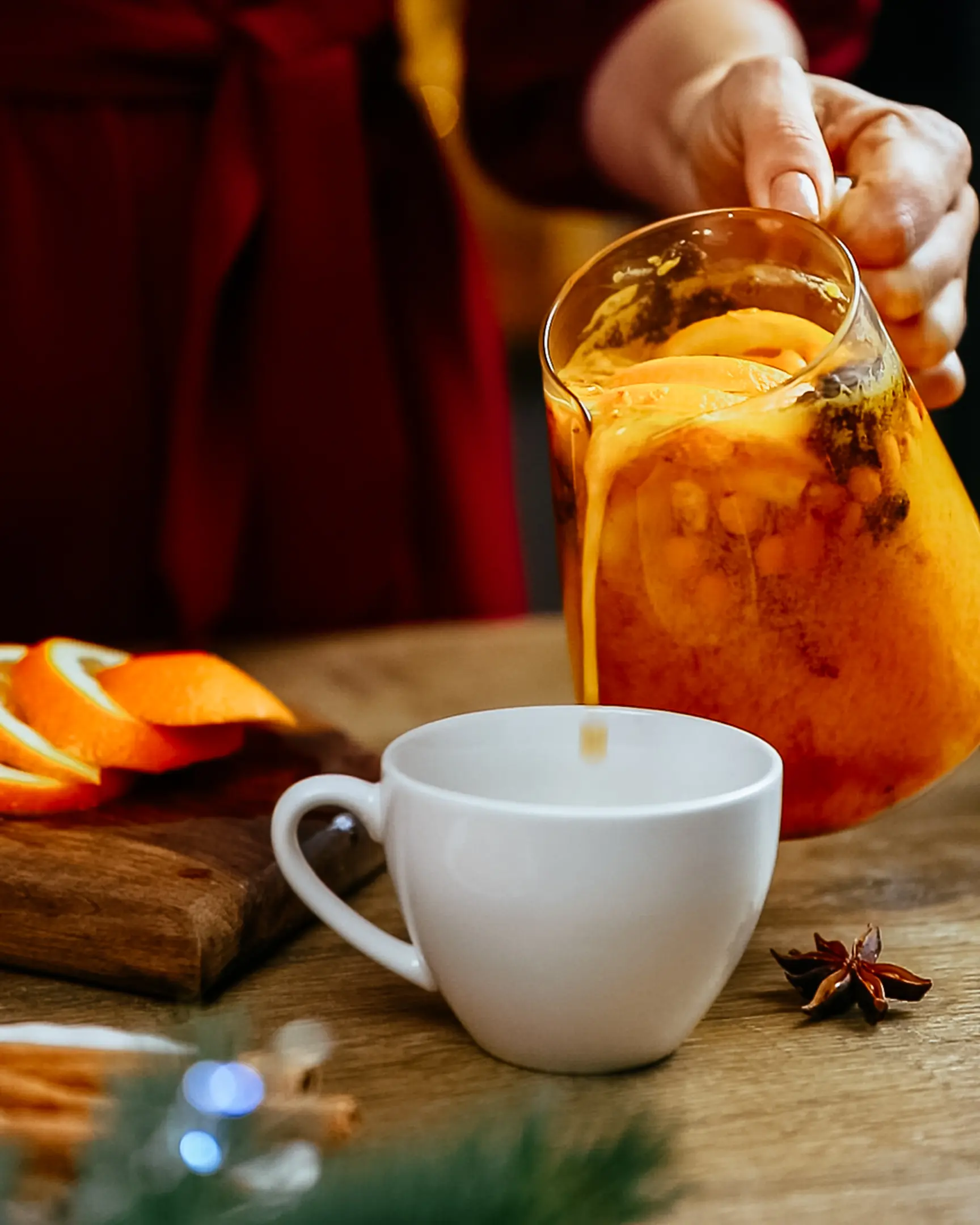 Hand pouring vibrant sea buckthorn & orange tea from pitcher into white mug, with star anise & cinnamon on table.