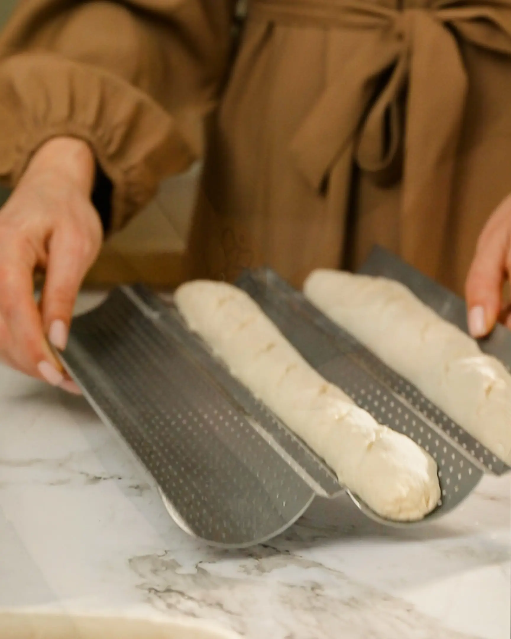 Shaping baguettes: Hands place formed sourdough or yeast dough into a perforated baking pan.